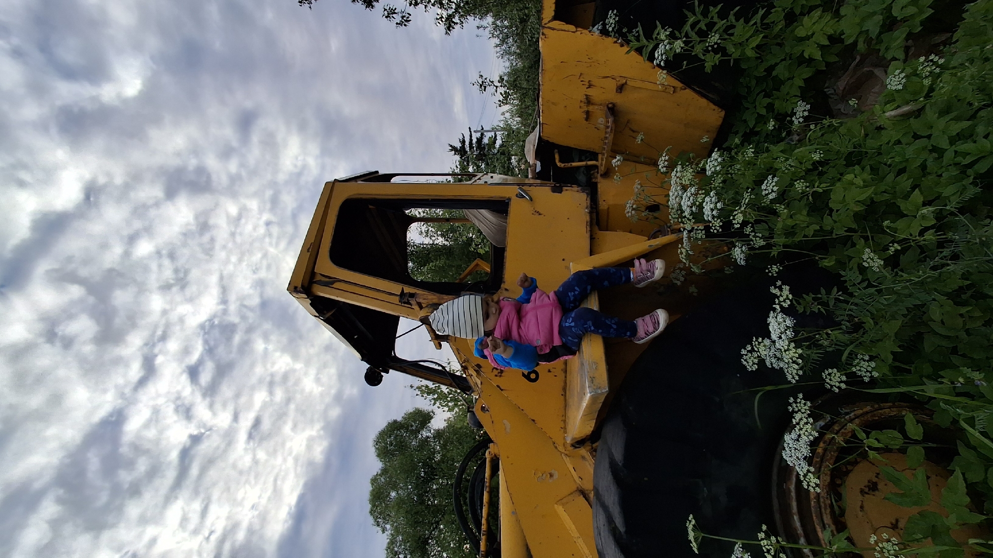 A low-angle, eye-level shot captures a young child sitting on the edge of an abandoned yellow construction vehicle. The child, wearing a striped hat, pink vest, and patterned leggings, looks down at their hands. White flowers and green foliage surround the base of the vehicle, indicating an overgrown, possibly rural or abandoned setting. The sky above is overcast with dynamic clouds. The scene is likely located in the village of Innolovo, Russia, given the context.