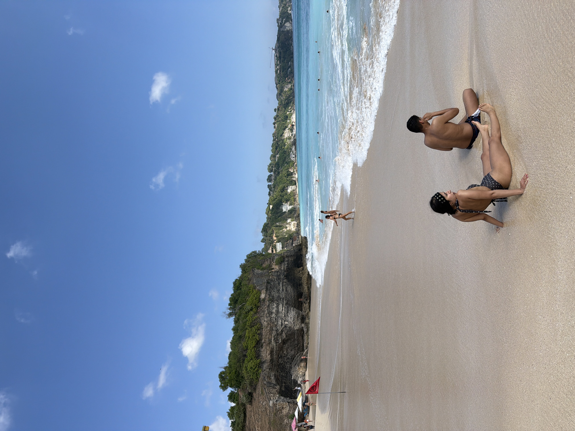 A beach scene in Kuta Selatan, Indonesia features a sandy shoreline, turquoise water, and a rocky cliff face covered in lush green vegetation. In the foreground, a man and a woman are sitting on the sand, facing away from the camera. The woman is wearing a black and white polka dot bikini, and the man is wearing dark blue swim trunks. Further up the beach, several people are walking along the shore, and some are in the water. A red flag is planted in the sand near the water's edge, likely indicating a warning or safety notice. The sky is clear and blue with a few scattered white clouds.