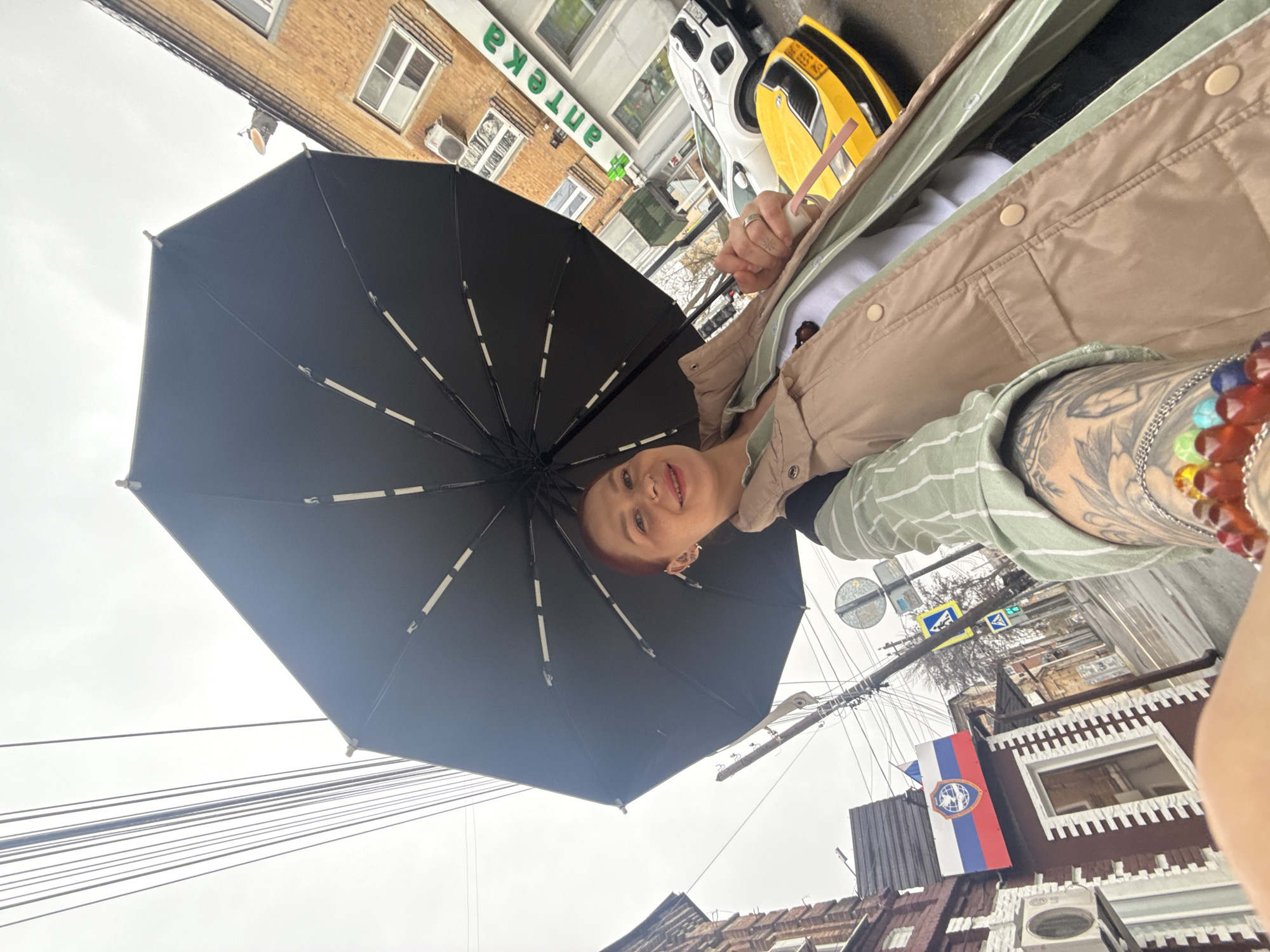 A low-angle selfie taken in Rostov-on-Don, Russia, features a woman holding a black umbrella. The background includes urban elements, specifically the “Apteka” sign (pharmacy in Russian), buildings, street signs, and a flag with a coat of arms against a tricolor background. A yellow and white vehicle is partially visible. The sky appears overcast. The woman has red hair, multiple ear piercings, and tattoos. She is wearing a beige jacket and striped shirt. She is holding a cup with a straw. The focus is primarily on her face and the umbrella.