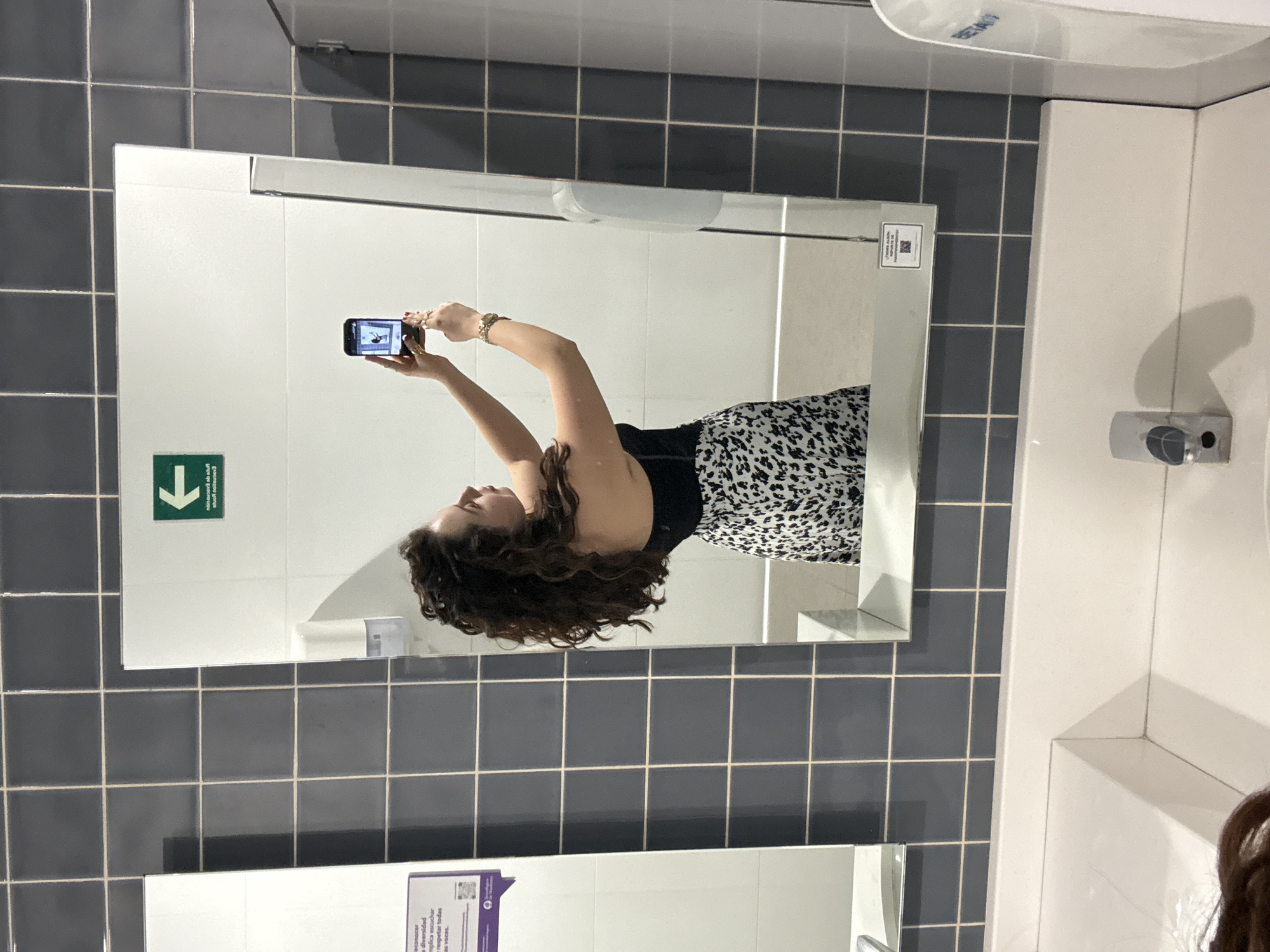A woman is taking a mirror selfie in a bathroom with gray tile walls and a white countertop. The reflection in the mirror shows the woman holding a phone in her raised hands, wearing a black top and a black and white spotted skirt. A green and white sign with a downward-pointing arrow is visible on the wall. A soap dispenser and a paper towel dispenser are on the wall above the countertop. Another mirror and a sign are visible in the bottom portion of the photograph. The location is Mexico City, Mexico.