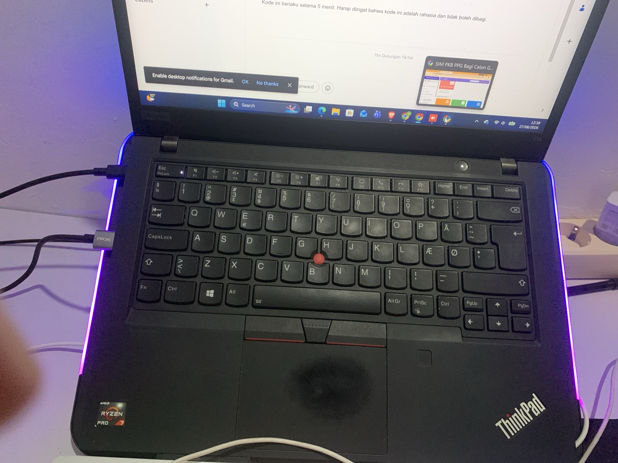 The image shows a close-up, high-angle view of a black Lenovo ThinkPad laptop open and positioned on a surface that appears to be a desk. The laptop is illuminated by a purple LED strip along its edge and ambient lighting. On the screen, there is a notification from Gmail asking to "Enable desktop notifications for Gmail" with options to "OK" or "No thanks." Below this, the Windows taskbar is visible, displaying icons for various applications including Google Chrome, Microsoft Edge, and file explorer. The timestamp on the taskbar indicates 12:39 on 27/08/2026. The keyboard layout is visible, showing standard QWERTY keys, along with function keys and navigation keys. The "ThinkPad" logo is prominently displayed on the lower right palm rest. The AMD Ryzen PRO 7 logo is visible on the lower left of the laptop's base. A portion of a person's hand is in the bottom left corner of the frame. Two cables are connected to the laptop's left side, one of which has a gray "PX" connector. The scene does not provide any direct visual cues to locate it specifically in Dukuh Mojo, Indonesia.