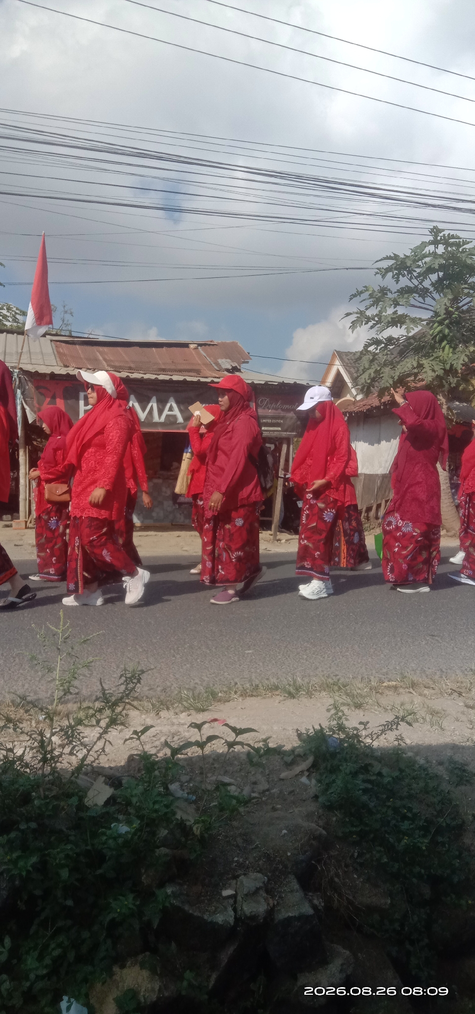 In Pakong, Indonesia, a group of people in red attire walks on a paved road, possibly participating in a parade or event. The individuals are dressed in matching outfits consisting of long-sleeved shirts and patterned skirts. Some are wearing headscarves, and a few have white caps or hats. They are walking in a line, facing towards the left of the frame. Behind them, a dilapidated building with a corrugated metal roof is visible, with a banner that appears to have "PAMA" written on it. A red and white flag is also present, planted on a pole near the building. The sky is overcast with some scattered clouds, and power lines are strung across the upper portion of the image. Lush green vegetation lines the side of the road in the foreground, with some rocks and debris scattered on the ground. The overall scene suggests a daytime event taking place in a somewhat rural or suburban area.