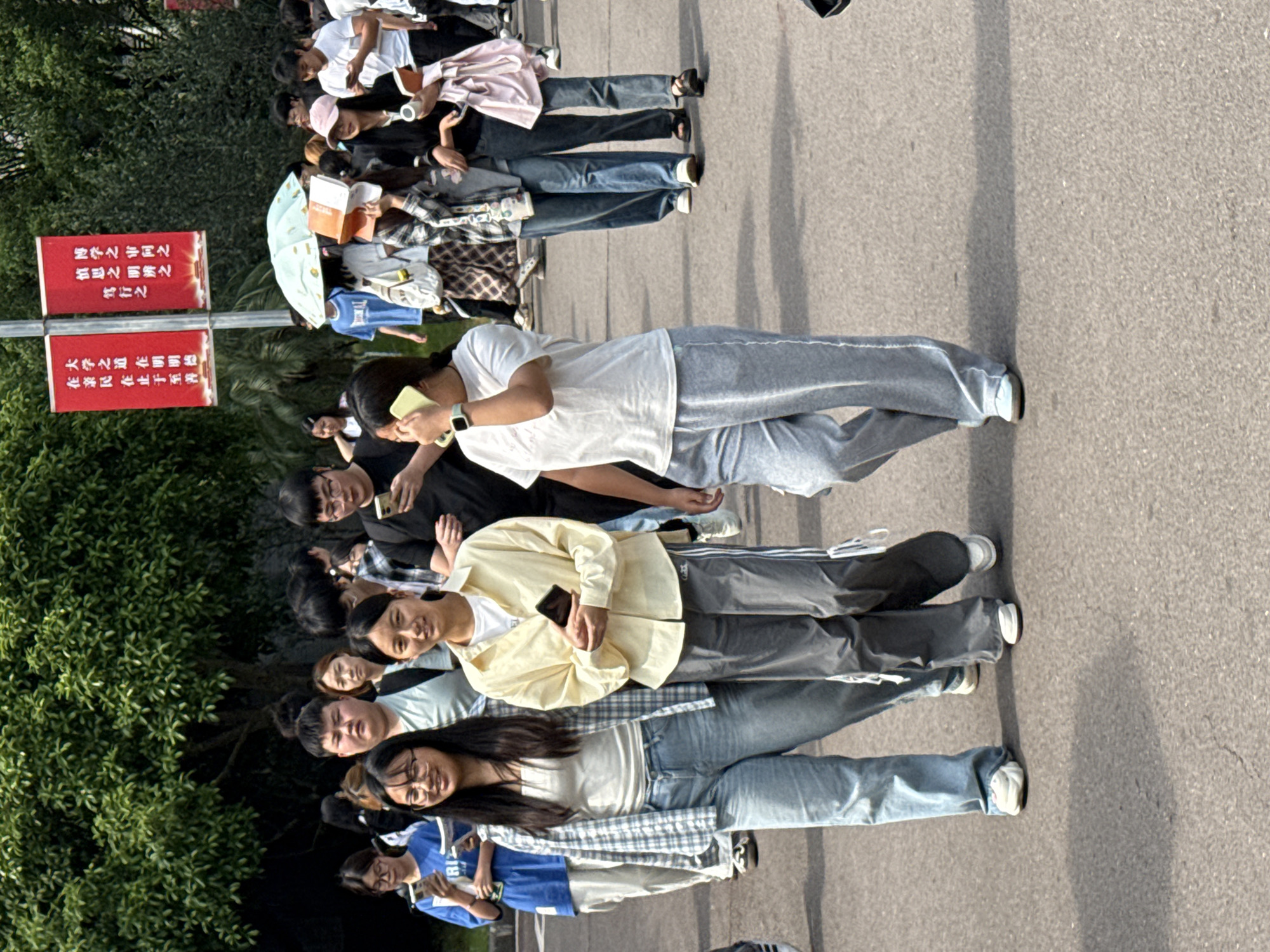 Here is a detailed description of the image:
**Location:** Jiangjin, China
**Scene:** A medium-shot perspective captures a group of people lining up on a paved area. Signposts are present, featuring text in Chinese characters.
**People:** Approximately 15 people are visible, primarily appearing to be young adults, possibly students. They are dressed in casual attire, including jeans, t-shirts, jackets, and athletic wear. Some are holding phones or books. The crowd is mostly facing toward the right.
**Objects:**
* Signposts: Two red signposts stand side-by-side, carrying Chinese text.
* Phones: Several individuals hold phones.
* Books: At least one person carries a book.
**Environment:**
* Paved ground: A paved surface serves as the backdrop.
* Vegetation: Green trees and foliage are seen.
* Signposts: Two red signposts stand side-by-side, carrying Chinese text.
**Actions and Interactions:** The people are gathered, likely waiting in line. Some are engaged with their phones, while others appear to be in conversation or reading.