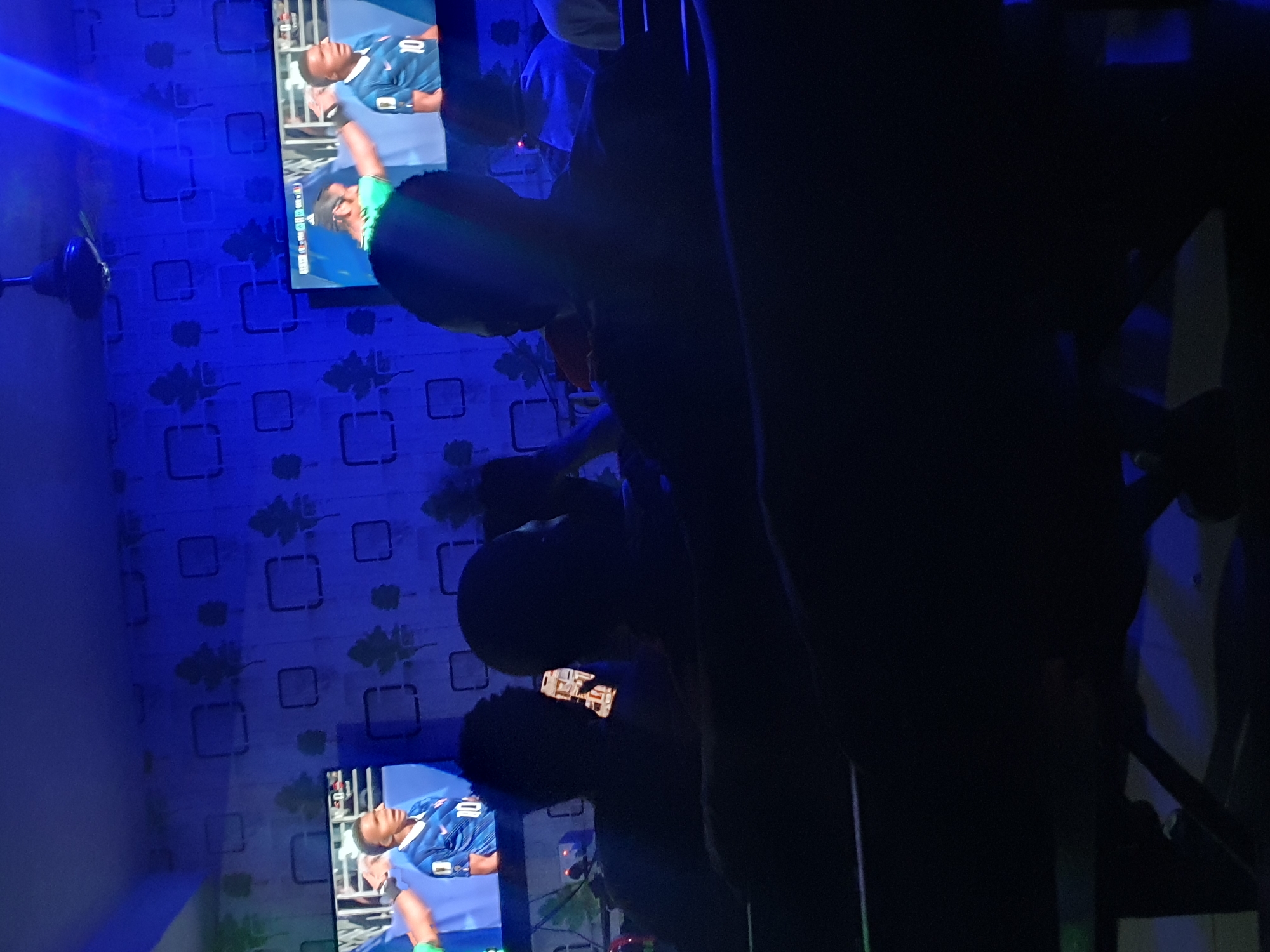 The image depicts a dimly lit room, possibly a lounge or viewing center, with a blue-patterned wallpaper. Two large screens are prominently displayed on the wall, showing a soccer match. On the left screen, a soccer player in a blue jersey with the number 10 is visible. The right screen also shows the same match, with a player in a blue jersey and another individual in a green jersey.
In the foreground, three individuals are silhouetted against the light from the screens. The person in the center is holding a smartphone, which is illuminated, suggesting they are interacting with it. The other two individuals appear to be watching the soccer match. The overall atmosphere is relaxed and casual, typical of a place where people gather to watch sports.
The scene is set in Lokoja, Nigeria, a city known for its vibrant culture and lively social scene. The viewing center could be a popular spot for locals to catch up on live sports events. The image captures a moment of shared enjoyment and community in a relaxed, informal setting.