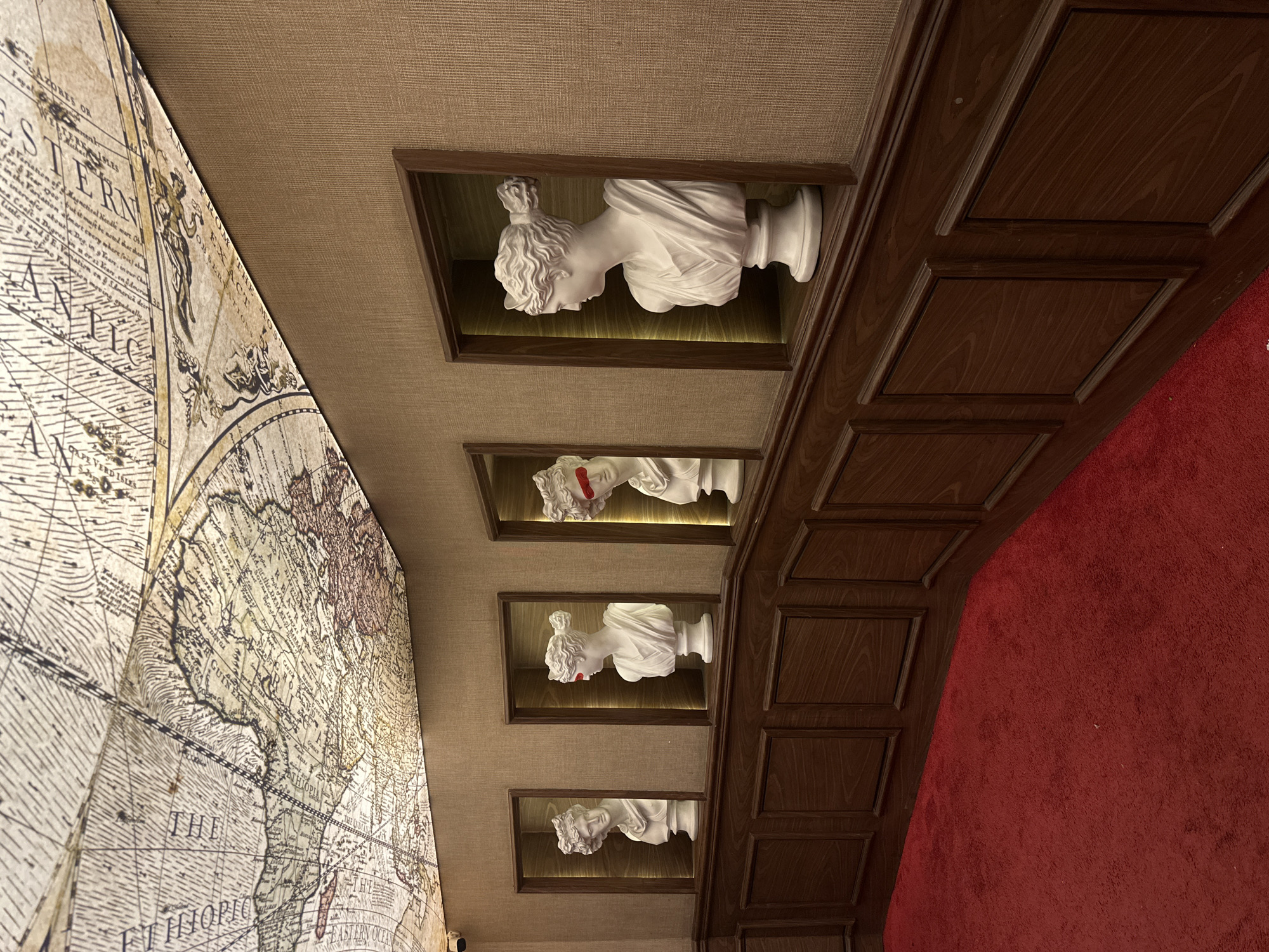 The image shows an interior view of a room. On the left is a wall covered with a large, old-fashioned map graphic. The ceiling is a light beige color. Along the top of the wall, recessed into the ceiling, are four white bust sculptures displayed in dark-wood framed niches with lighting. The busts are spaced in a vertical row. A dark wood wall with decorative panels runs along the right side, bordering a red carpeted area. The photo appears to have been taken from a low angle, pointing upwards. The location is likely not Itirin, Nigeria, based on the image content.