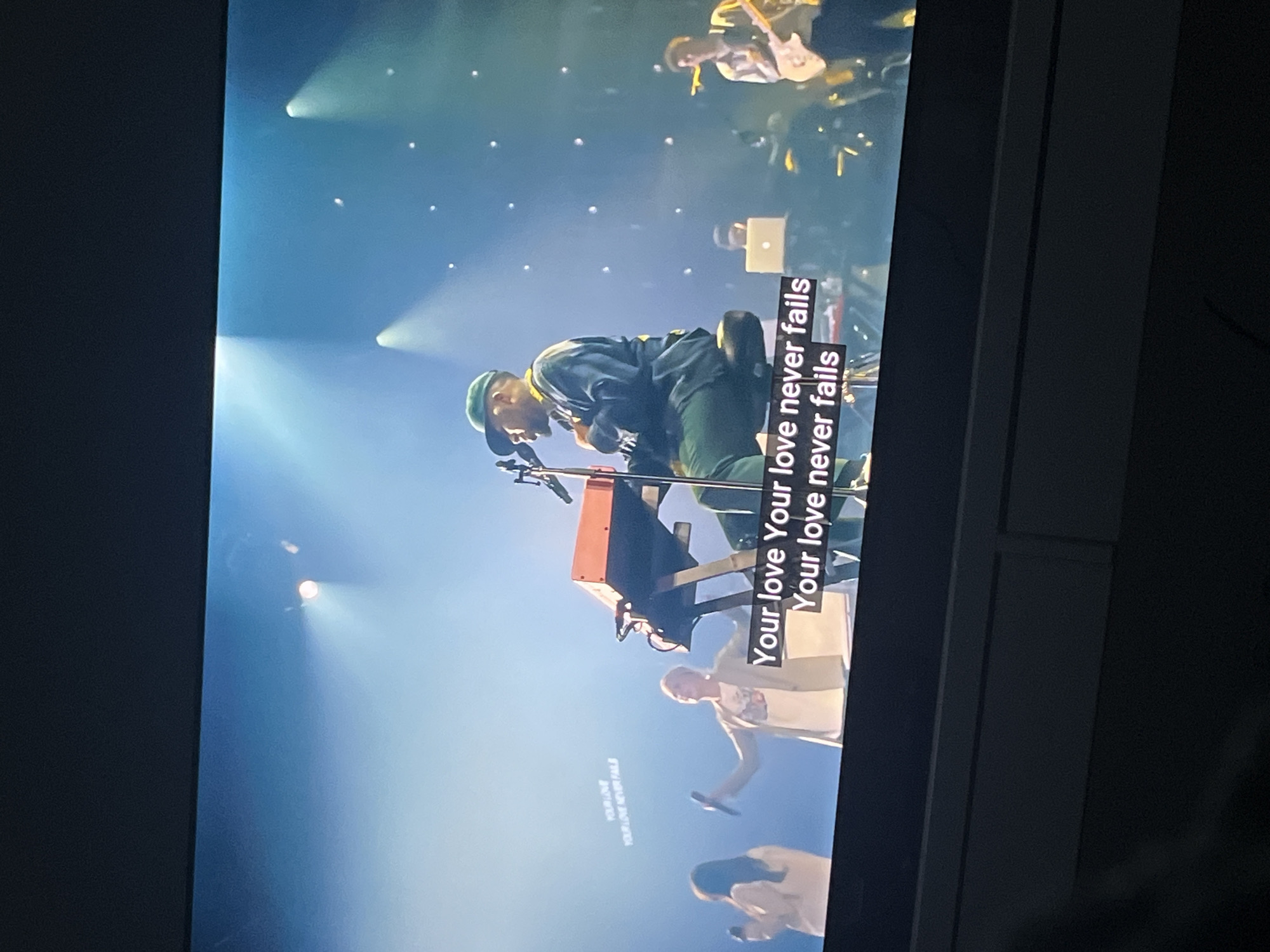 The image shows a television screen displaying a concert performance. A musician in a green hat is seen playing a keyboard center frame, with the lyrics "Your love Your love never fails" superimposed on the screen. Several other performers are visible in the background, including singers holding microphones and a guitarist. The lighting is bright, creating a stage-like atmosphere. The location, Mogán, Spain, is not directly evident from the visual information in the image.