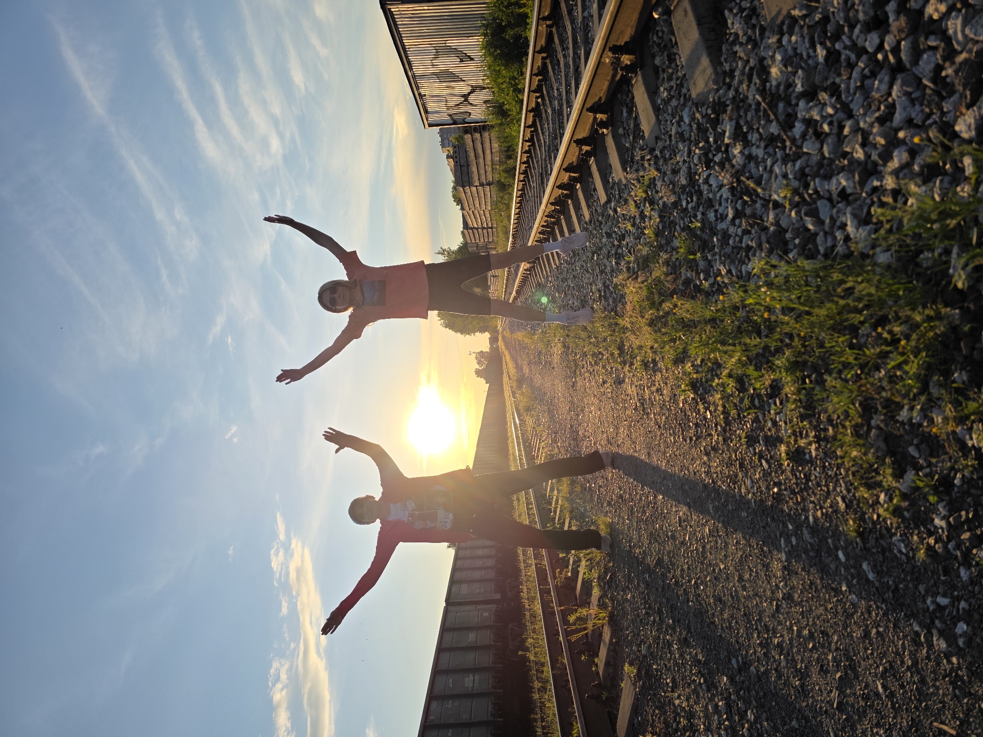 Two individuals are captured in mid-air, jumping on a railway track under a bright, partly cloudy sky during sunset. The person on the left wears a red jacket over a graphic t-shirt and dark pants, with arms outstretched upwards. The person on the right, wearing sunglasses, a coral-colored t-shirt, and dark shorts with white socks and sneakers, also has arms raised. Both individuals are silhouetted against the setting sun, which casts long shadows across the gravelly tracks and surrounding vegetation. In the background, freight train cars are parked on adjacent tracks, and a long line of stacked concrete barriers runs alongside the tracks, suggesting an industrial or logistical area. The overall scene depicts a moment of joy and freedom in a somewhat industrial urban landscape. The location is the railway sidings near the Kama River in Chaikovsky, Russia.