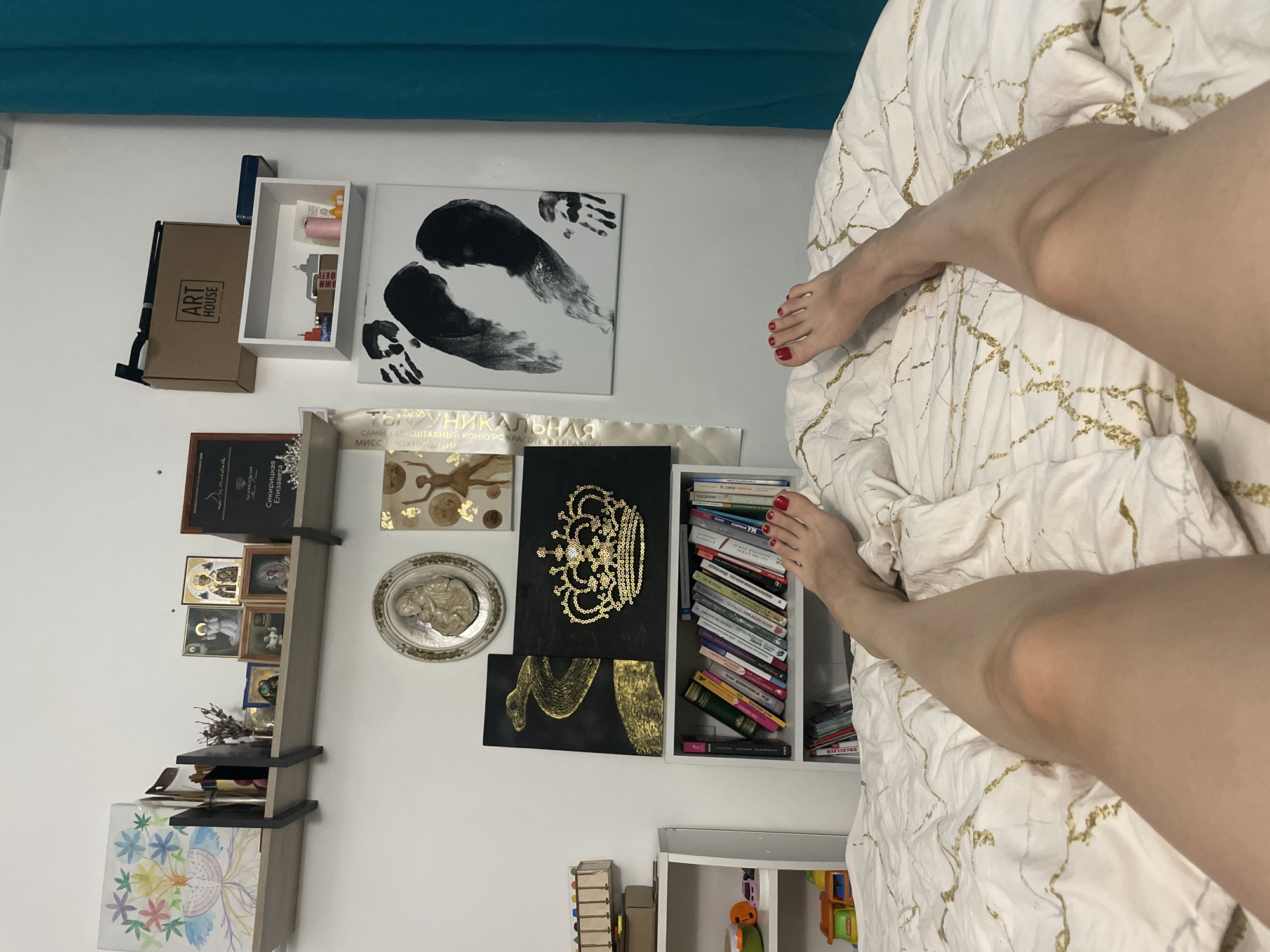 This indoor scene features an unusual perspective, seemingly taken from above while lying on a bed. The bed is covered with a white and gold patterned duvet. In the foreground are two legs, positioned in a V-shape. The toes are painted a vivid red.
The background wall is painted white and decorated with various artworks and shelves. A painting with black handprints is visible, along with a shelf displaying smaller framed pictures and decorative objects. There is also a vertical shelf crammed with books. Artwork featuring gold paint and a black backdrop can also be seen. The upper part of a teal curtain can be seen at the top of the frame.