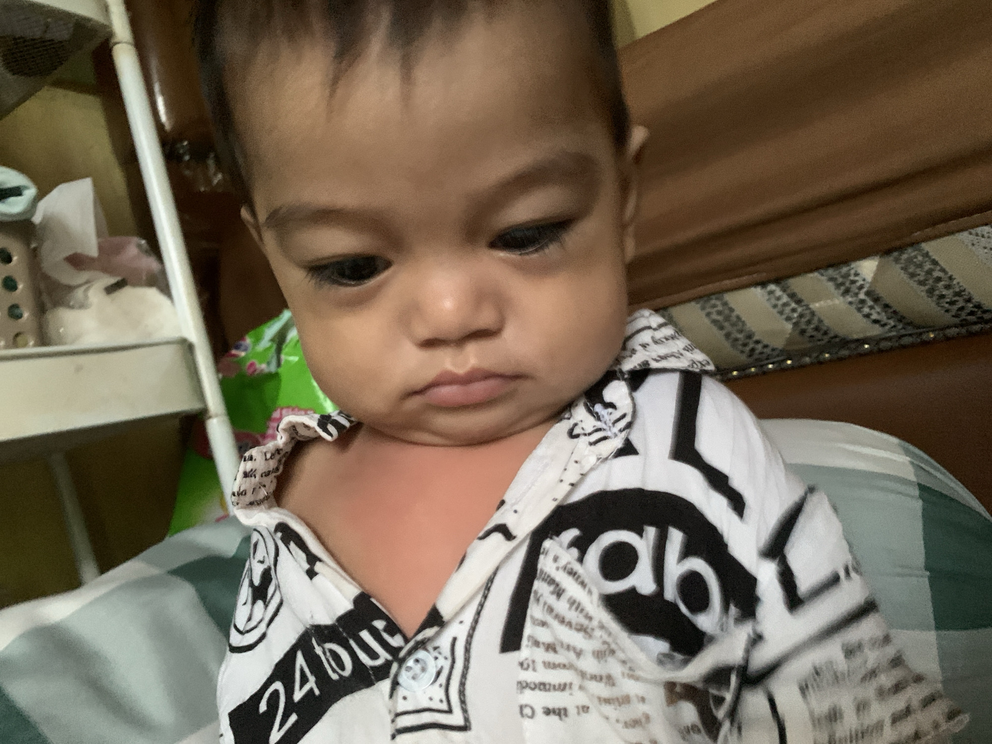 A close-up, medium-shot photograph shows an infant in Sungai Meriam, Indonesia. The infant has brown hair and brown eyes and is wearing a white shirt with black lettering. The shirt is partially unbuttoned, exposing the infant's chest. The infant is looking down with a slightly pouty expression. In the background, there is a wooden headboard of a bed. A metal utility cart with tissues and other items is in the left background. The bedding is a green and white plaid pattern.