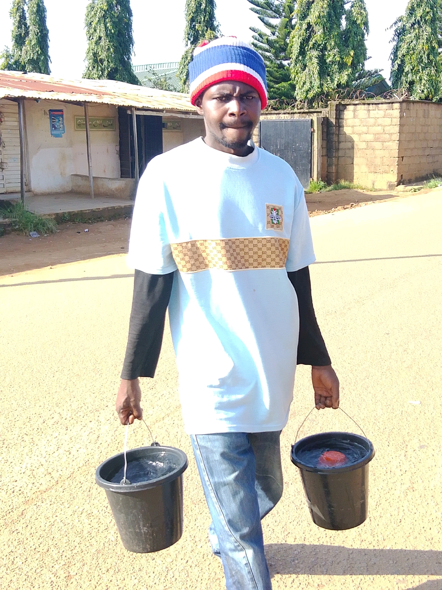 A young Black man wearing a blue, white, and red striped beanie, a light blue t-shirt with a decorative gold band and bee emblem, and blue jeans walks towards the camera carrying two black buckets of water. One bucket is held in his left hand, the other in his right. The scene appears to be a dirt road in a residential area of Jos, Nigeria. In the background, there is a single-story building with a corrugated metal roof, a gate, and tall green trees. The sky is clear and blue.