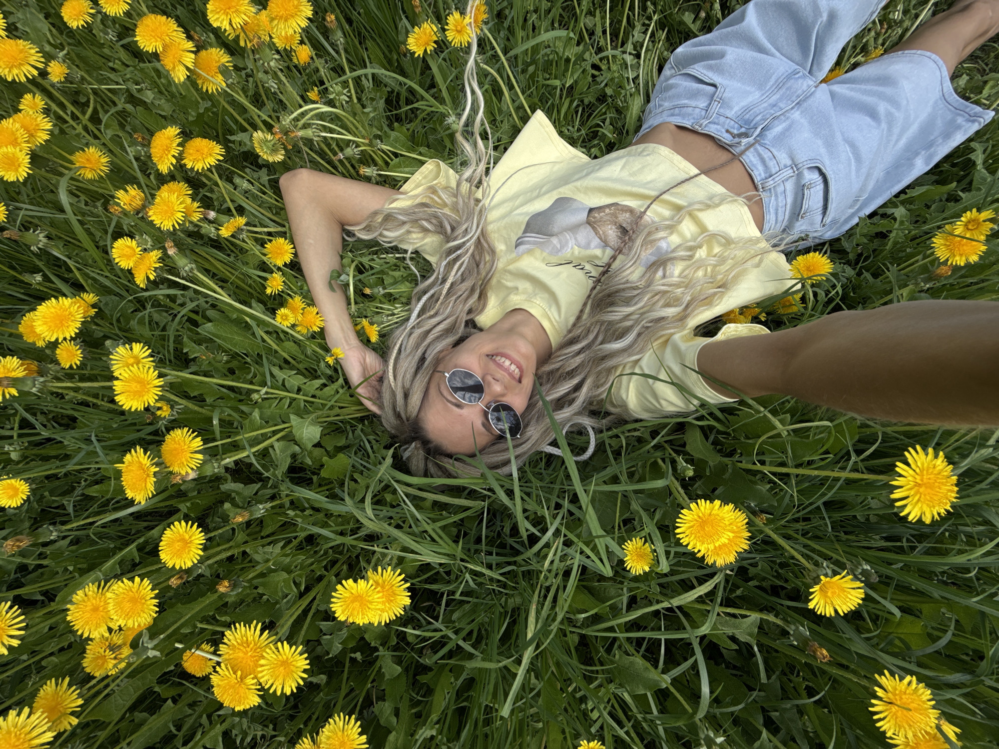 A woman lies in a field of dandelions, with her head towards the bottom of the image and her feet towards the top. She is wearing light blue jeans, a yellow t-shirt, and sunglasses. Her blonde hair is styled in waves. The field is densely covered with green grass and bright yellow dandelions. The scene appears to be outdoors. The location is Unknown, Unknown.