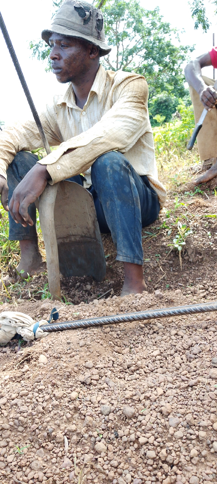 The image depicts a close-up view of a man, likely a construction worker, sitting on the ground. He is wearing a faded, patched bucket hat, a light-colored, long-sleeved collared shirt, and dark blue jeans. His hands rest on a shovel, and his bare feet are visible in the dirt. The ground is covered in soil and small rocks. In the foreground, a thick metal rod, possibly rebar, lies on the ground, partially covered by a white cloth bundle tied with a blue string. In the background, another person, only partially visible and wearing a red shirt, is also present, as well as green vegetation and trees, suggesting an outdoor environment. The scene appears to be located in Kamantan, Nigeria, a town known for its iron ore deposits and related mining activities.