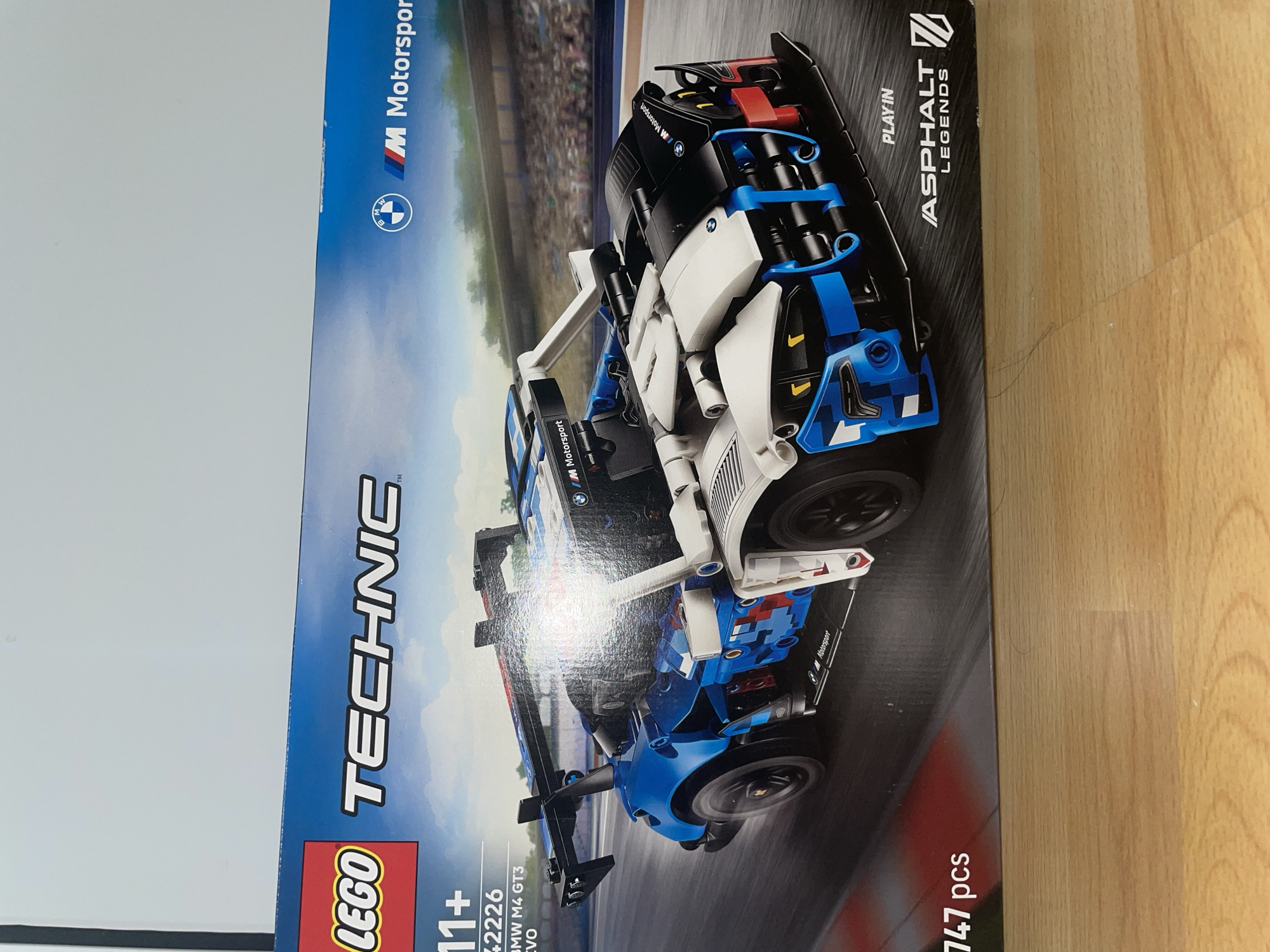 The image shows a LEGO Technic box. The box is for the 42226 BMW M4 GT3 EVO model, with a recommended age of 11+. It is a Lego Technique and the LEGO logo can be seen in the bottom left hand corner. The set contains 747 pieces. The box features the BMW M Motorsport logo and indicates compatibility with the Asphalt Legends game. The background of the box shows a blurred image of a racetrack. The box is placed on a wooden surface.