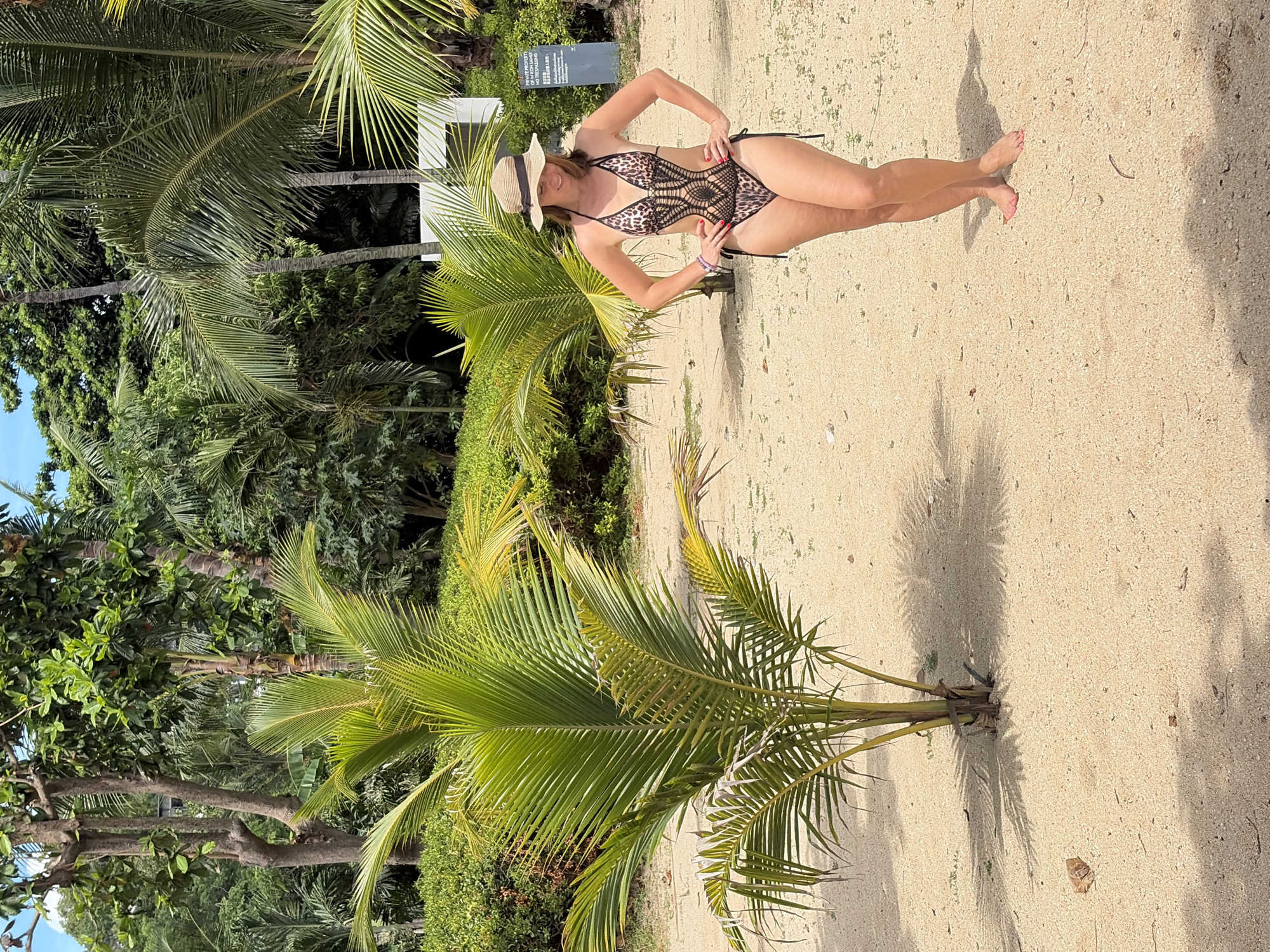 A woman stands on a sandy beach in Baan Bo Phut, Thailand, wearing a leopard-print and black crochet one-piece swimsuit and a straw hat. She has her hands on her hips and is looking down. A small palm tree is positioned to her left, and a lush green hedge is behind her. In the background, there are more palm trees and dense foliage, typical of a tropical environment. To the right, a white sign with black text is visible, indicating "PRIVATE PROPERTY NO TRESPASSING." The sand is light brown and shows shadows from the palm trees. The sky is bright and partly cloudy.