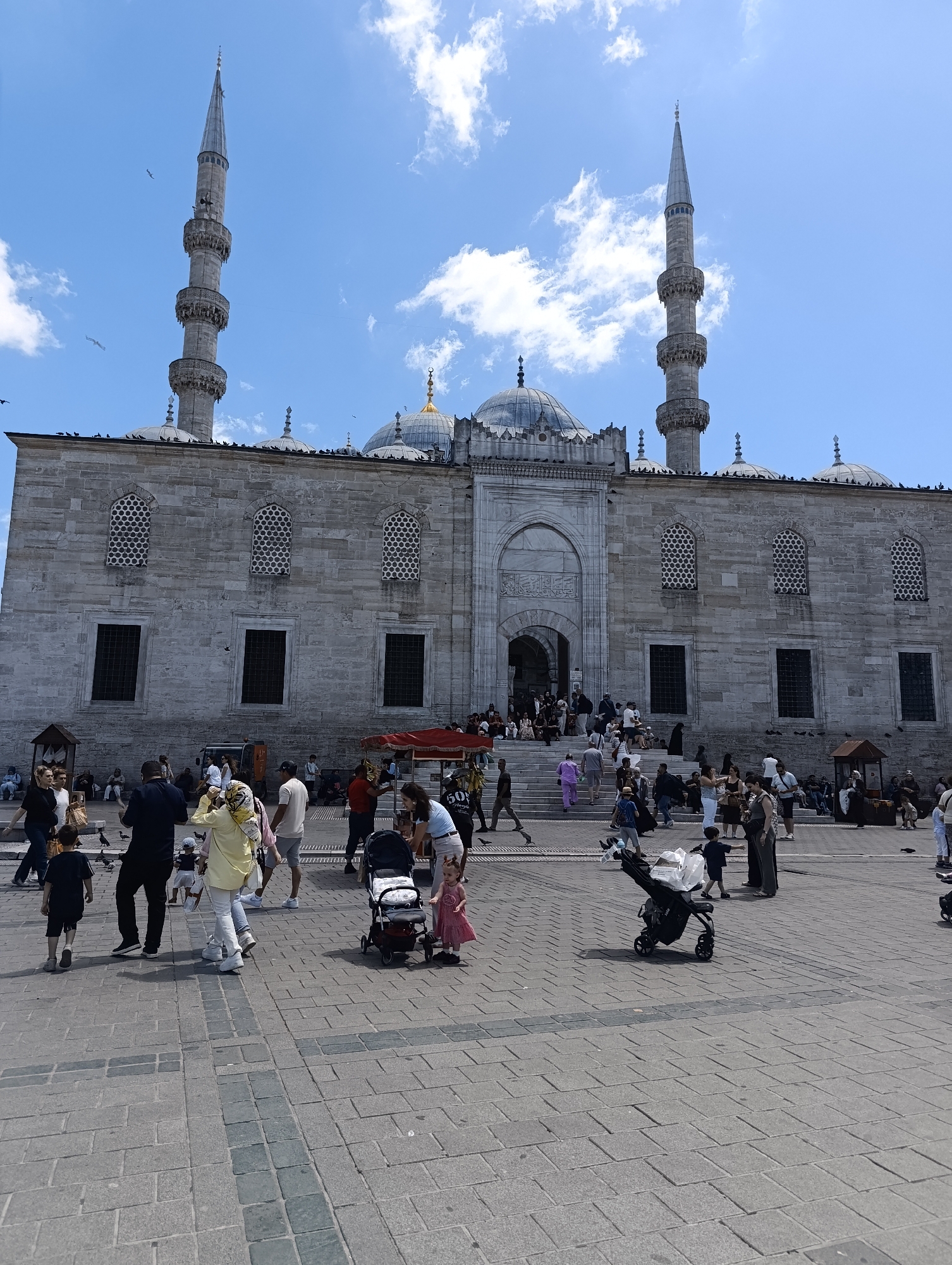 The image captures a view of the Süleymaniye Mosque in Istanbul, Turkey, on a sunny day with a partly cloudy sky. The imposing mosque features a grand stone facade, multiple domes, and two tall, slender minarets. A large, paved courtyard in front of the mosque is bustling with activity. Numerous people are walking, standing, and sitting, creating a vibrant atmosphere. Among them are individuals pushing strollers, suggesting families are visiting. Some people are dressed in casual attire, while others wear more traditional clothing, including headscarves. Birds are visible perched on the mosque's roof and flying in the sky, adding to the dynamic scene. The ground is paved with large, light-colored stone tiles, some of which are arranged in a patterned inlay. Small kiosks or stands are present in the courtyard, hinting at vendors selling goods or services. The overall impression is one of a popular tourist destination and an active public space.