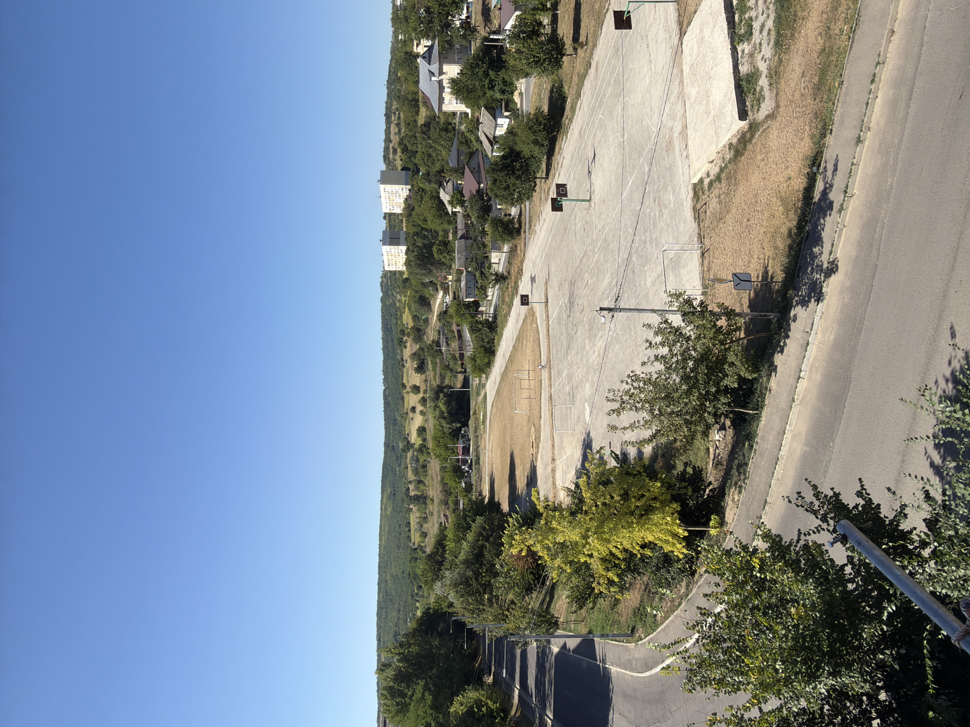 This is a high-angle, full shot of a town in Strășeni, Moldova, on a clear, sunny day. The scene is dominated by a large, empty, outdoor concrete basketball court with several hoops. Adjacent to the basketball court is a grass field with what appears to be a soccer goal. A paved road curves through the foreground, bordered by a sidewalk and green trees. In the middle ground, there are several residential buildings of varying sizes, including a few multi-story apartment buildings in the distance. The background is filled with rolling, tree-covered hills. The image appears to be taken from a building overlooking the area, capturing a wide expanse of the town and its surroundings. There is no visible activity or people in the image.