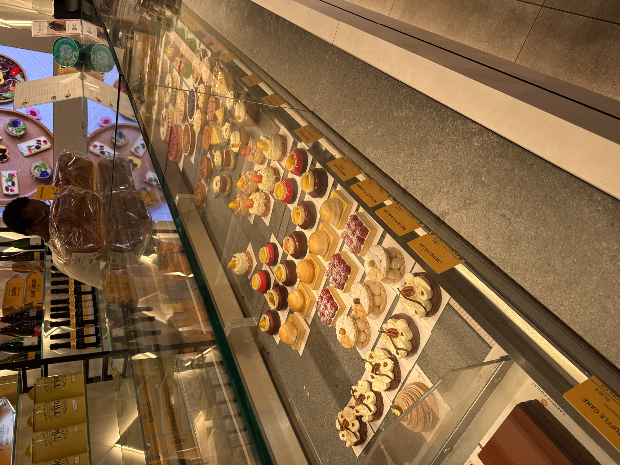 Here's a detailed description of the image:
**Scene:** The image shows a refrigerated display case filled with a variety of pastries, cakes, and other delicacies. The display appears to be inside a shop, possibly a bakery or patisserie.
**Subjects/Objects:**
* **Pastries/Cakes:** An extensive assortment of individual pastries and cakes, including tarts, mousse cakes, and layered desserts. Many are decorated with fruit, cream, or nuts.
* **Labels:** Small yellow labels are placed next to many of the pastries, presumably indicating names and prices. Some of the labels are visible, showing prices in Euros, and names like "DACQUOIS MAISON", "TARTELETTE FRAMBOISE", "CROUSTI PÉCAN".
* **Other Products:** In the background, shelves are visible with bottles of wine, jars of what appears to be tea, and loaves of bread.
* **Glass Display:** The pastries are displayed behind a clear glass case, which is part of the refrigerated unit.
* **Person:** A person is visible in the reflection of the glass, wearing a face mask.
* **Environment:** The display case is clean and well-lit. The background suggests a high-end food shop.
* **City:** Labels like “REIMS-75440M” and mentions of “KAEMPEE-KOHLER” suggest that this is a branch of the German brand “KAEMPFF-KOHLER”. The bottles also include "Saint Amour" and "Capri."
**Actions/Interactions:**
* The pastries are on display, likely for sale to customers.
**Location (Based on the information available):**
Based on the "KAEMPFF-KOHLER" brand affiliation, it is likely that the shop is located in the city of Luxembourg, Luxembourg.