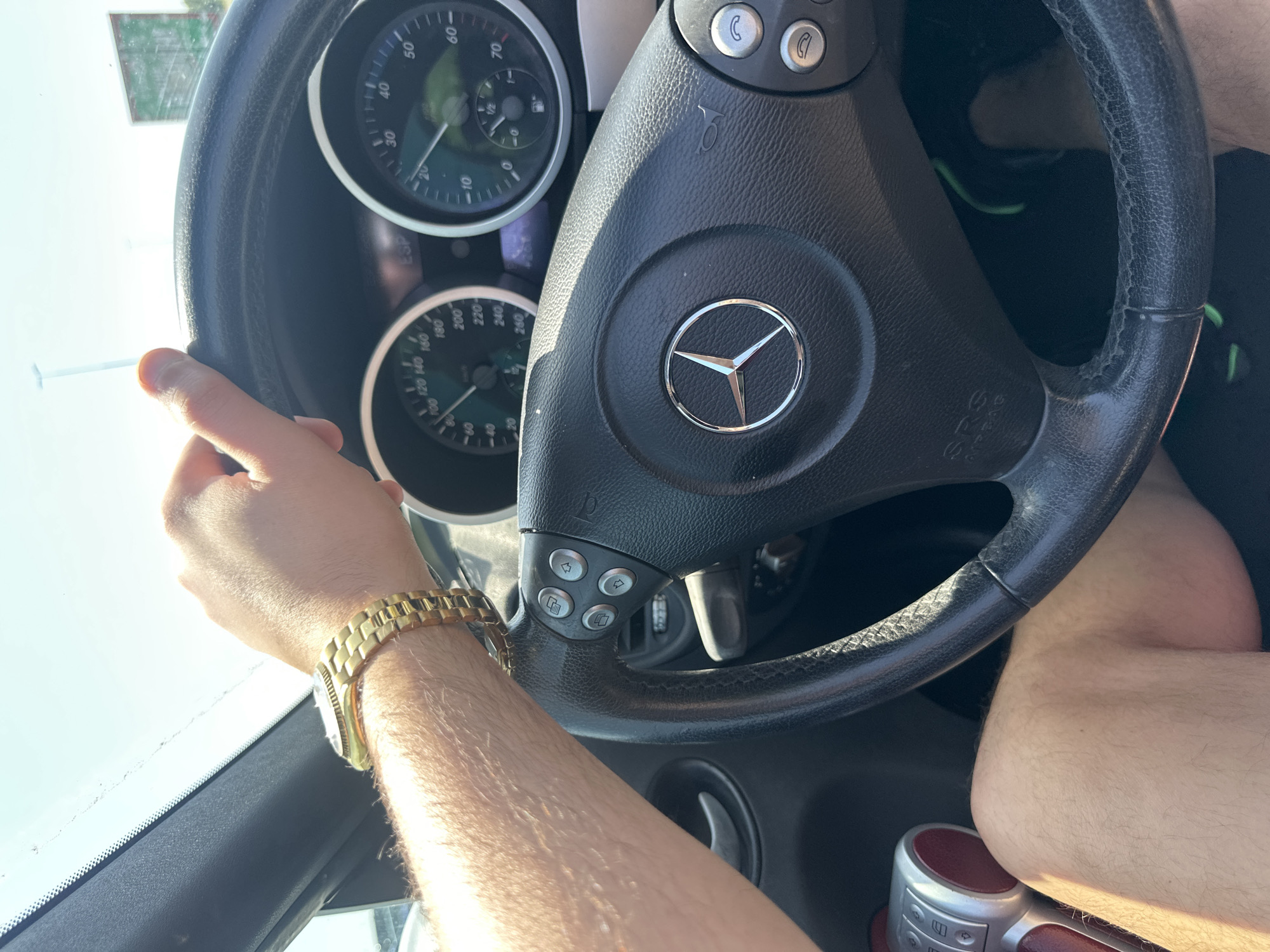 The image shows the interior of a Mercedes-Benz car. The dashboard is visible, with the steering wheel featuring the Mercedes-Benz logo prominently displayed. The speedometer and fuel gauge are partially visible behind the steering wheel. A male hand with a gold watch is gripping the left side of the steering wheel. Part of the man's legs are also visible. There is no information to determine that this image was taken in Leidschendam, Netherlands.