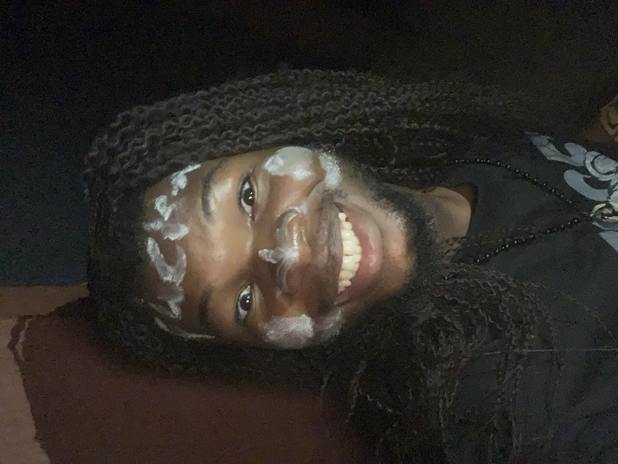 A person with dark skin and long, dark, curly hair is smiling at the camera with a white substance smeared on their forehead, nose, and chin. The person is wearing a black shirt and a black beaded necklace. The background is dark and blurry. The photo is taken at an angle with the person's head tilted to the side. There is no indication of the location being Onitsha, Nigeria.