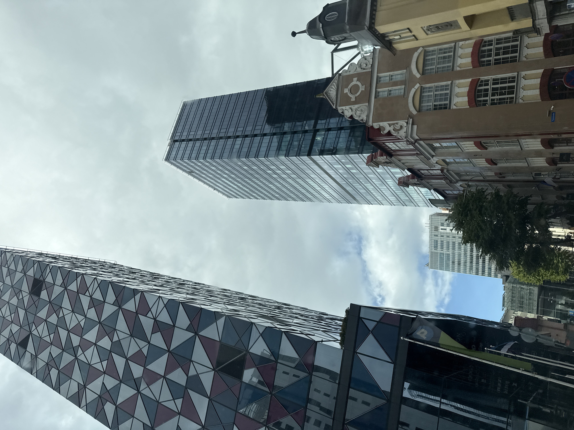 This image shows a view of Tallinn, Estonia, looking up at several buildings. In the foreground, on the left, is a modern, tall building with a facade of geometric triangular patterns in shades of gray, purple, and blue. To the right of this, and slightly behind it, is another very tall, modern skyscraper with a dark, reflective glass facade. In front of the glass skyscraper and to the right, there are several older, ornate buildings with traditional European architecture, featuring pitched roofs, multiple windows, and decorative elements. One of these older buildings is light brown with red trim and accents, and another adjacent building is yellow. The sky above is overcast with some patches of lighter clouds. In the mid-ground, a group of trees can be seen, and further back, another, shorter modern building with a lighter facade is visible. Two flags are visible on poles in front of the geometric building on the left.