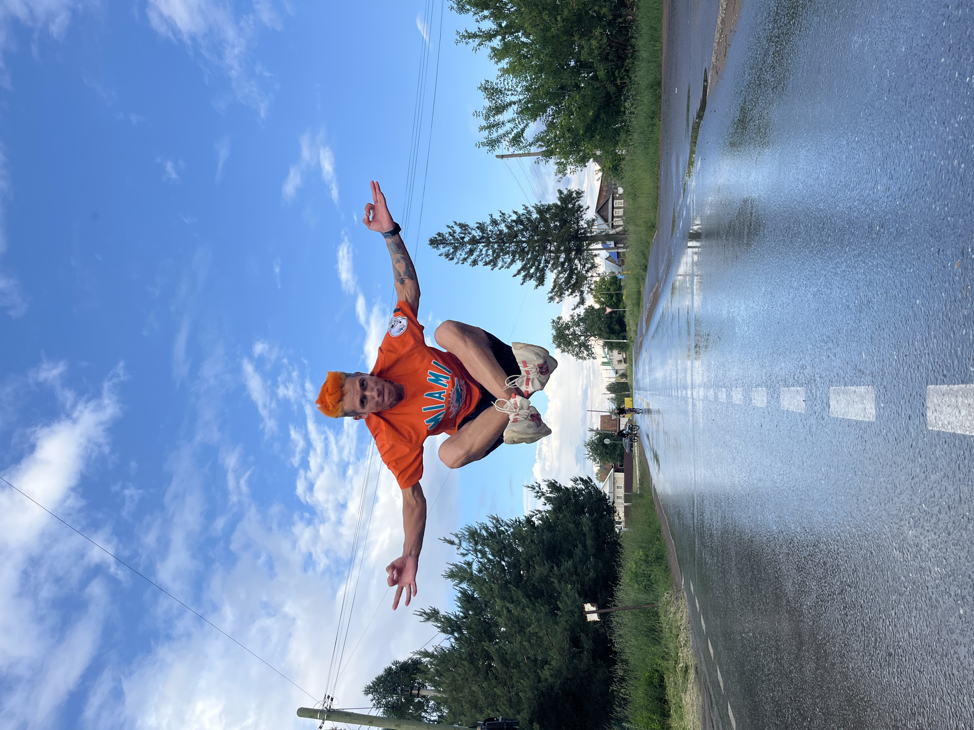 An action shot captures a Caucasian male mid-air, jumping above a wet, rural road. He has bright orange hair and is wearing an orange t-shirt with the word "MIAMI" and a logo, black shorts, and white sneakers with red laces. His arms are extended, and his hands are positioned in a mudra gesture, with his left hand forming a circle with his thumb and index finger. The road, marked with white dashed lines, reflects the cloudy blue sky and surrounding greenery. Trees line the sides of the road, and in the background, several houses and two people on bicycles are visible, suggesting a quiet village setting. The overall impression is one of dynamic movement against a backdrop of everyday rural life.