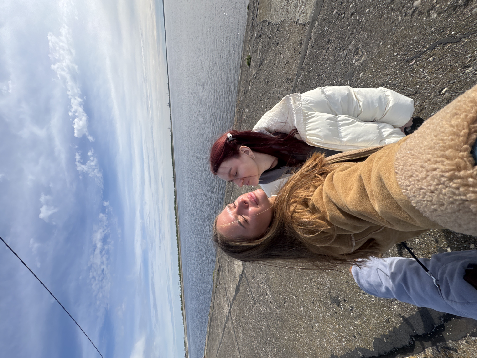 Two women are posing for a selfie outdoors on a concrete surface. One woman has long brown hair and is wearing a brown jacket. The other woman has red hair and is wearing a white puffer jacket. Behind them is a body of water and a cloudy sky. A power line is visible in the sky. Based on the location information provided (Ivanovo, Timiryazevskii S/S, Russia), it is possible that the body of water is the Volga River or one of its tributaries.