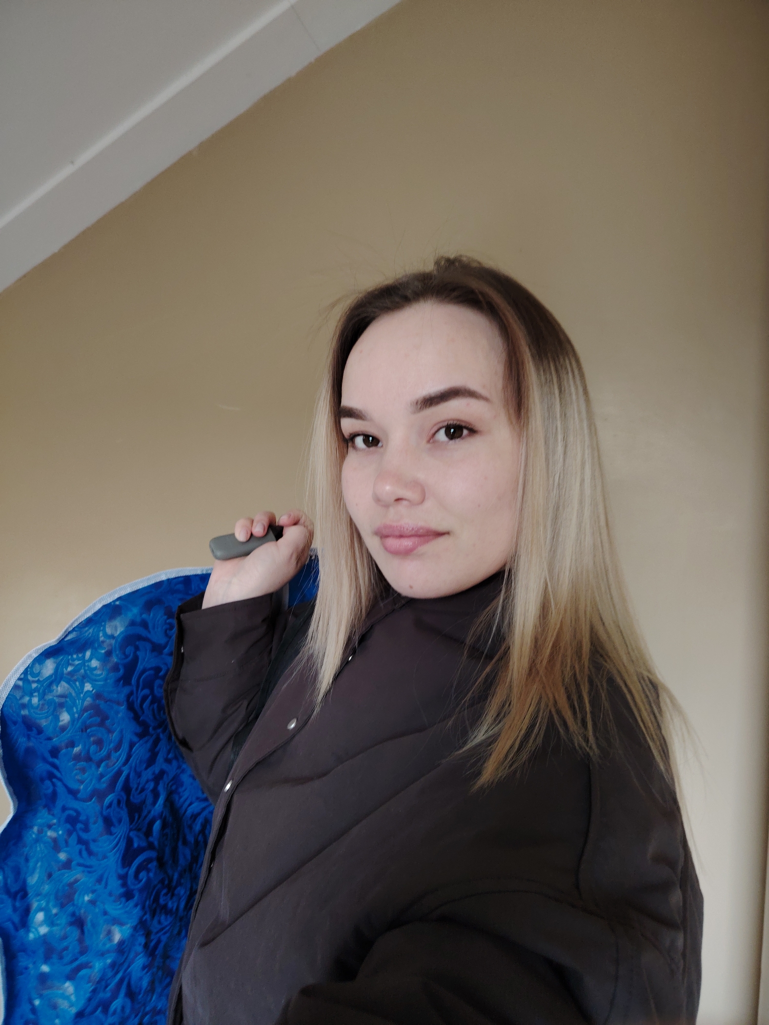 A head-and-shoulders portrait captures a young woman looking directly at the camera with a subtle smile. She has fair skin, dark brown eyes, and long, straight hair that transitions from darker roots to lighter blonde ends, parted on her left side. Her lips are a natural pink and slightly parted. She is wearing a dark, possibly black or dark grey, puffer jacket, which is partially visible. Her right hand, raised near her shoulder, holds a small, dark grey, unidentifiable object with a rounded shape. On the left side of the frame, behind her, a vibrant royal blue fabric or material with an intricate, swirling damask-like pattern is visible. The background is a plain, light beige wall, with a white ceiling or molding visible in the upper left corner, suggesting an indoor setting. The lighting appears soft and even, indicating daytime. There is no visible text in the image.