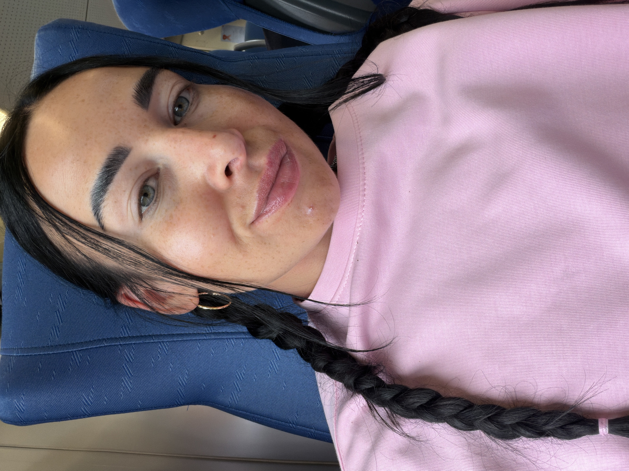 In the city of Ушаки, Russia, a young woman with dark hair, styled in two braids, is captured in a close-up selfie. She is seated on a blue textured chair, likely in a public transport setting, given the visible headrest and seat fabric. The woman is wearing a pink long-sleeved shirt. Her facial features are the focal point, with her eyes, eyebrows, and lips clearly visible. She is wearing gold hoop earrings. A small blemish is noticeable on her chin. The lighting suggests an indoor environment, possibly during the day.