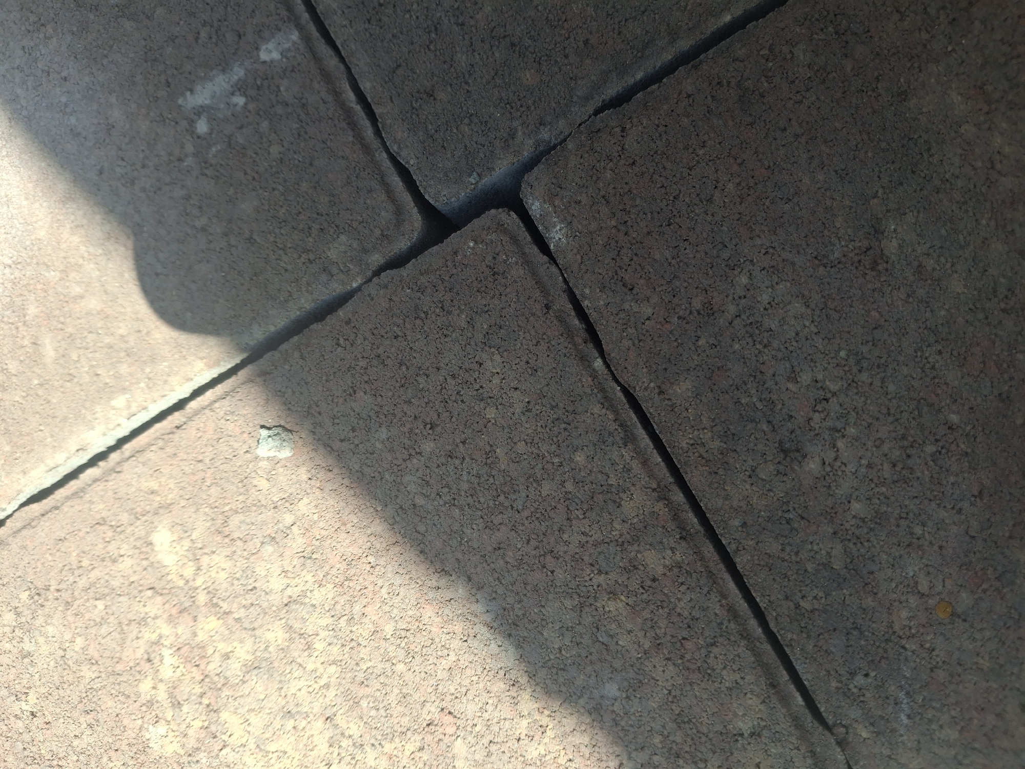 The image shows a close-up view of interlocking pavers, likely forming a sidewalk or patio. The pavers are textured and appear to be made of concrete or a similar material, with visible aggregate. They are a reddish-brown and dark grey color. A shadow of what appears to be a person's head and shoulder is cast diagonally across the upper right portion of the image. There is a small, light-colored piece of debris or a small stone on one of the lighter pavers. The lighting suggests it is daytime, with sunlight creating the shadow and illuminating parts of the pavers. The scene is an outdoor urban or suburban environment. The location of Kalush, Ukraine, is not discernible from the visual information in the image itself.