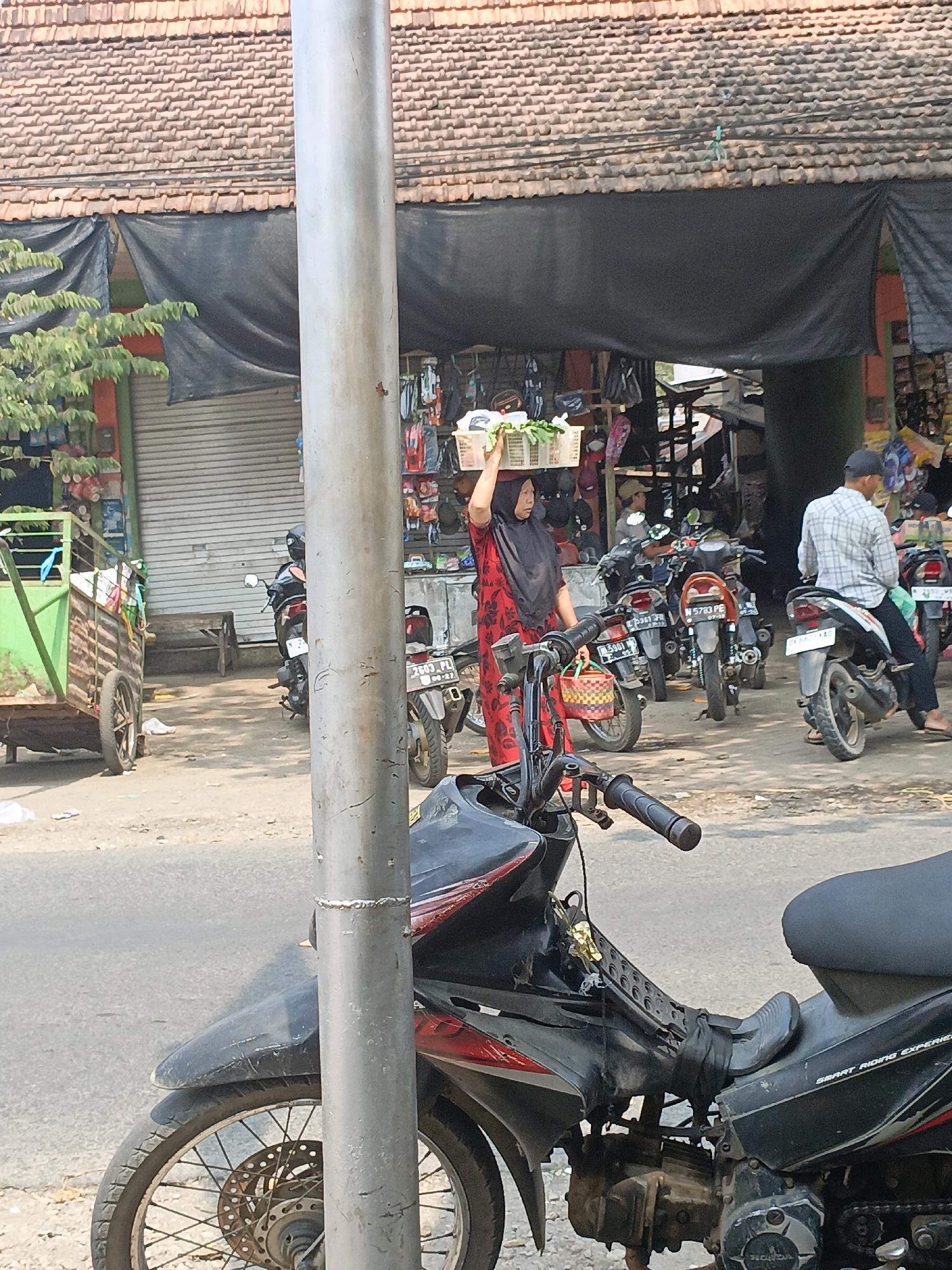 A street scene in Betek, Indonesia, features a woman wearing a red and black patterned dress and a black hijab carrying a basket of produce on her head. She is walking past a row of shops with their fronts covered by tarps and metal rolling doors. Several motorcycles are parked along the street, and a man in a plaid shirt is seated on one of them. In the foreground, a black motorcycle is partially visible, and a silver pole stands in front of the shops. The ground is paved with asphalt and dirt.