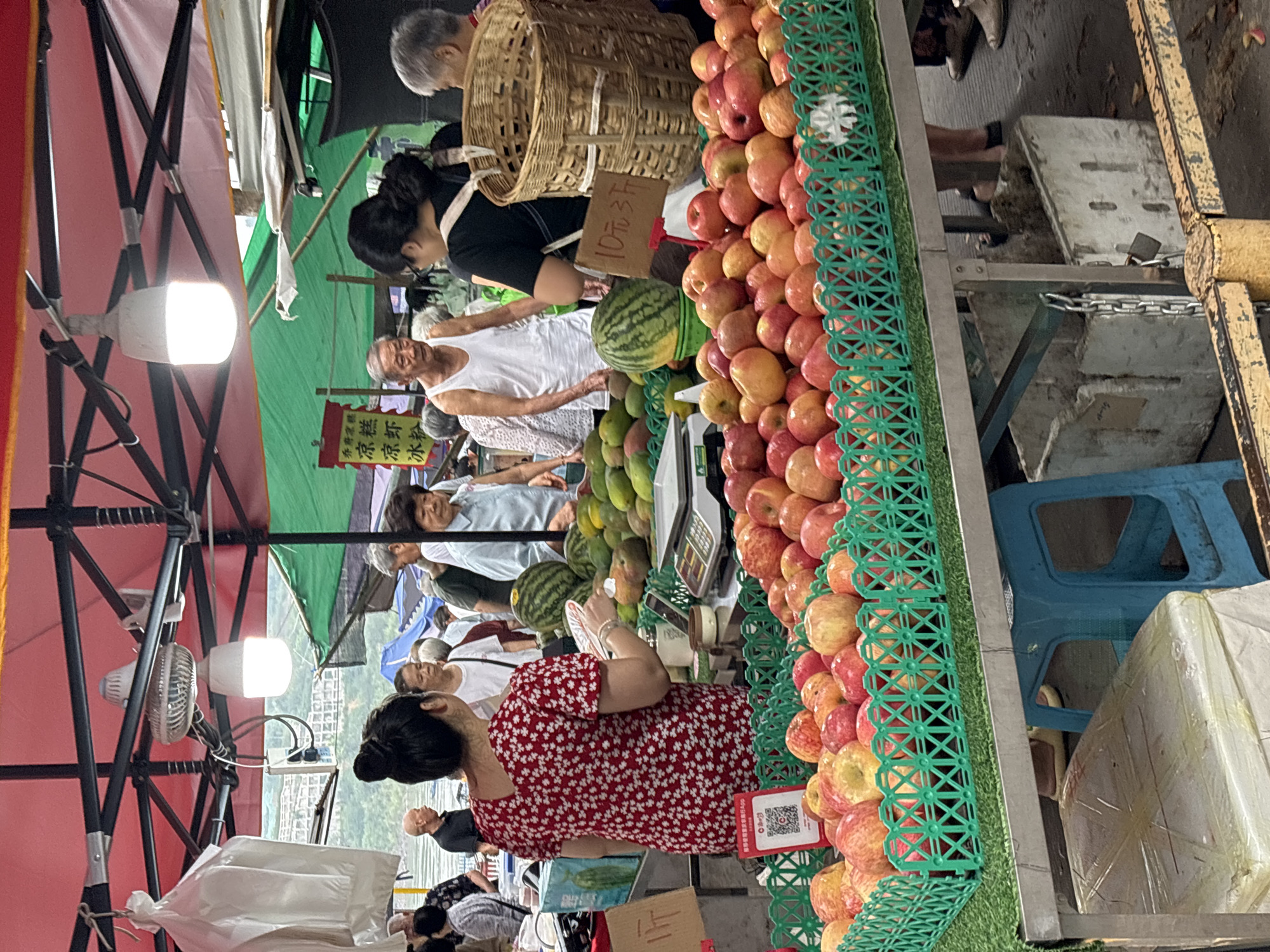 An outdoor fruit stall in Jiangjin, China displays an abundance of fresh produce for sale. A woman in a red floral dress is interacting with customers at the stall, which is laden with green crates overflowing with red apples. Behind the apples, a variety of other fruits, including watermelons and mangoes, are visible. Customers are browsing the selection, and a vendor can be seen weighing produce. The stall is covered by a red awning, and market activity is ongoing.