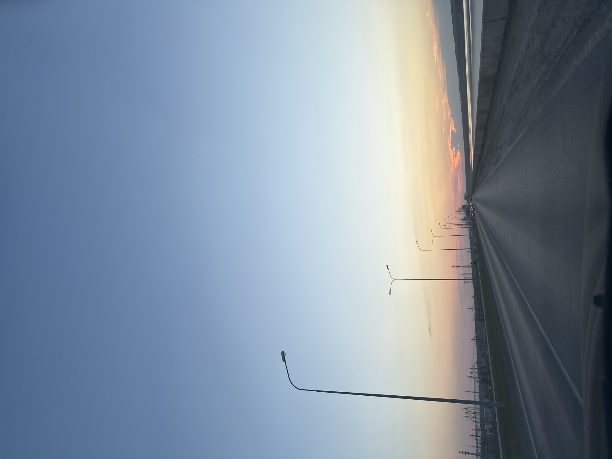The image depicts a highway lined with streetlights and power pylons, receding into a sunset on the edge of the city of Chaikovsky, Russia. The perspective suggests the photo was taken from inside a moving vehicle. The sky transitions from a deep blue to pale yellow where it meets the horizon. A concrete barrier runs along the right edge of the highway. There are a few cars visible in the distance.