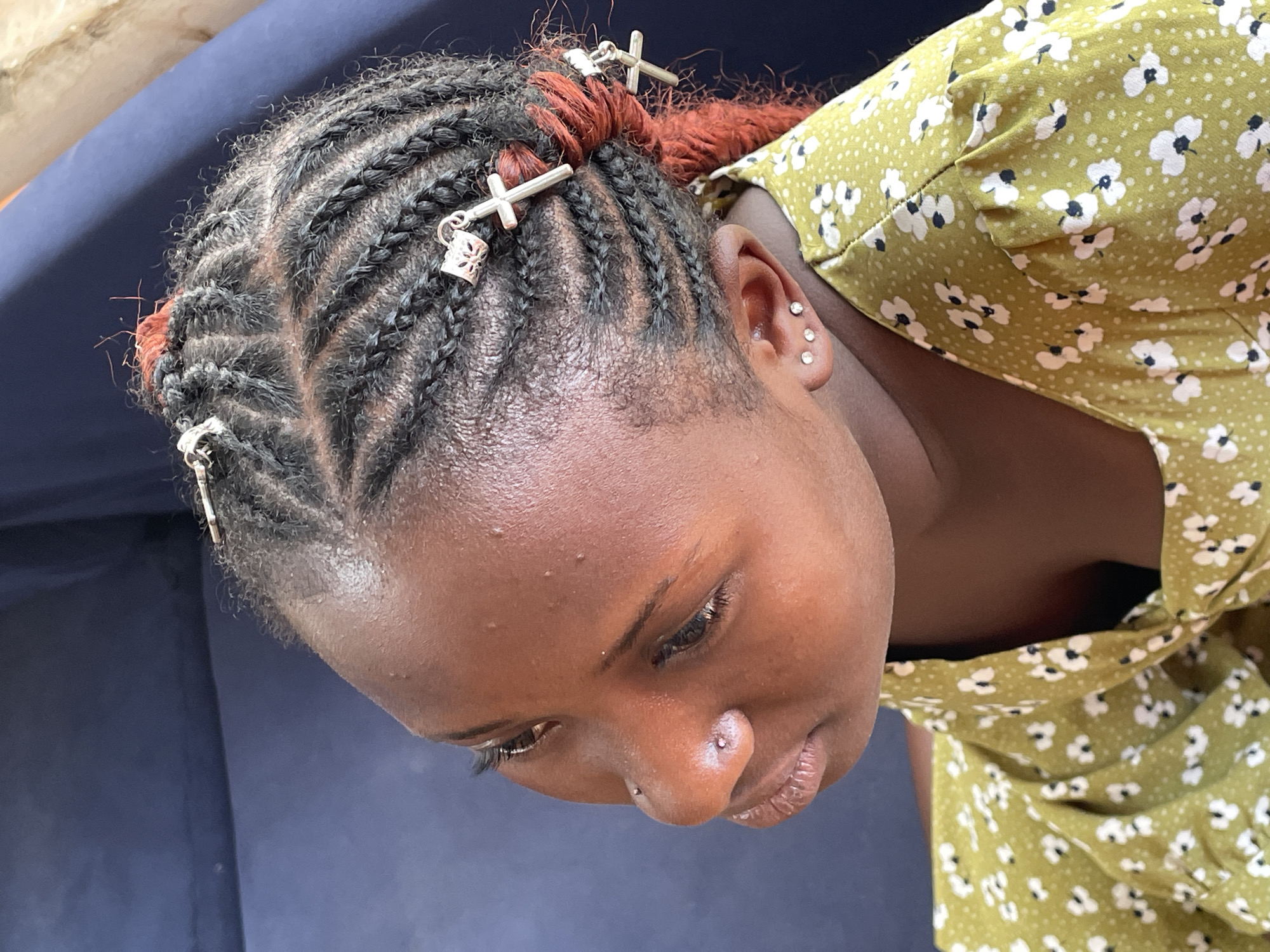 An overhead close-up shot captures a young person with dark skin, likely in Barakin Akawo, Nigeria. The subject's head is tilted downwards, eyes looking slightly to the right, giving a contemplative or shy expression. Her hair is intricately styled in cornrows, with some sections incorporating reddish-brown hair extensions. Several silver hair accessories, including cross-shaped charms and patterned beads, are woven into the braids.
The individual has multiple piercings: a small stud in her left nostril and three small, sparkly studs on her right earlobe/helix. Her skin has a natural sheen, possibly from sweat or natural oils, and a few minor blemishes are visible on her forehead. She is wearing a light green garment adorned with a pattern of small white flowers and dark centers, possibly a dress or a blouse with gathered fabric around the shoulder. The background is a solid dark blue or purple surface, suggesting an indoor setting or a shaded outdoor area. No specific activity is taking place; the person appears to be in a relaxed pose. The lighting is soft and natural, indicating it is likely daytime. There is no visible text in the image.