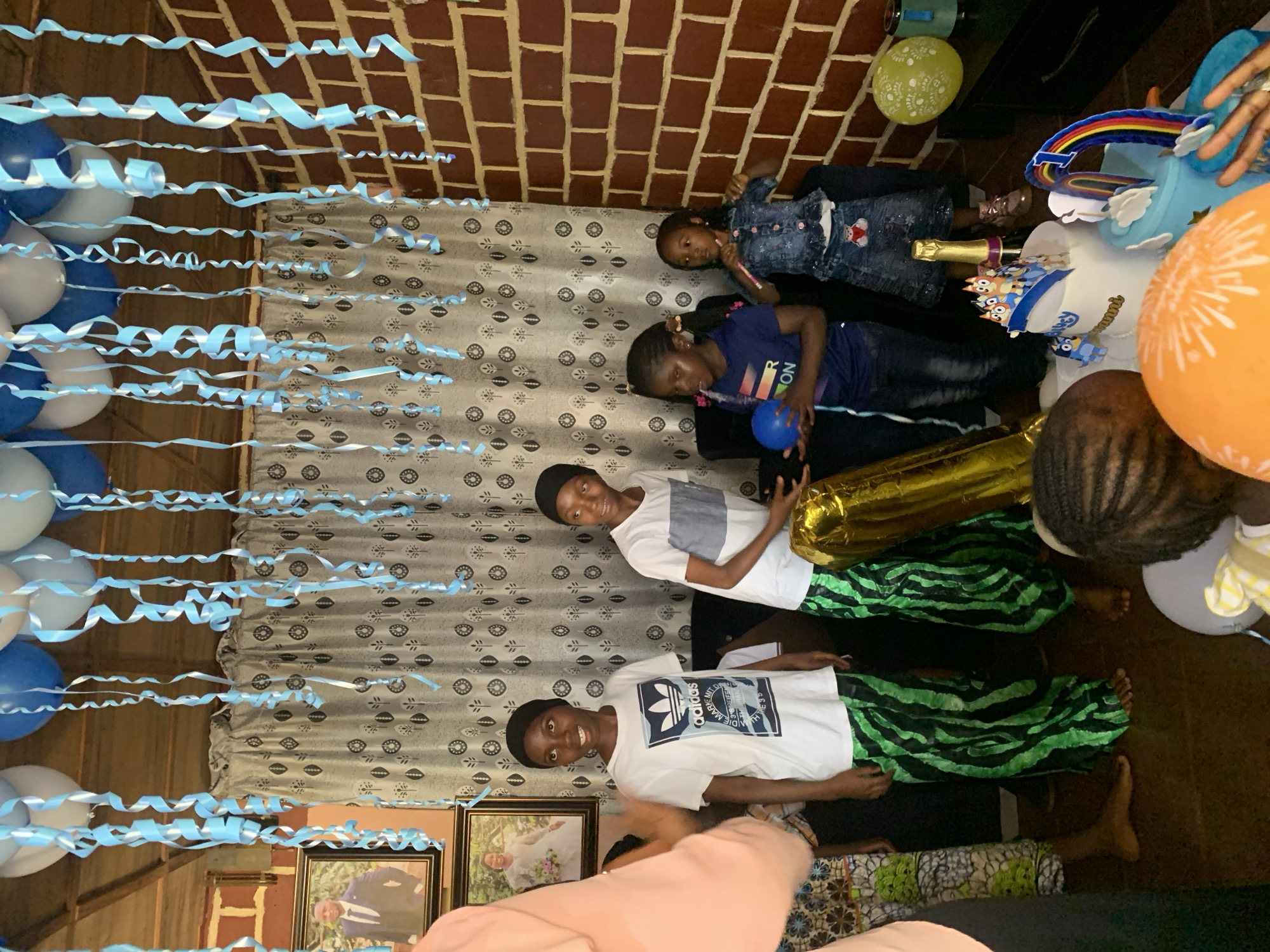 The image captures an indoor scene, seemingly a birthday celebration with a group of people in Jos, Nigeria. There are four children visible. The children are lying on the floor. Decorations are hung from the ceiling. There are balloons and cake. The floor is tiled in brown. There are 2 framed photos on the bottom left corner. There are light blue streamers along the top of the shot and light blue, white and dark blue balloons on the left side. The wall is brick.