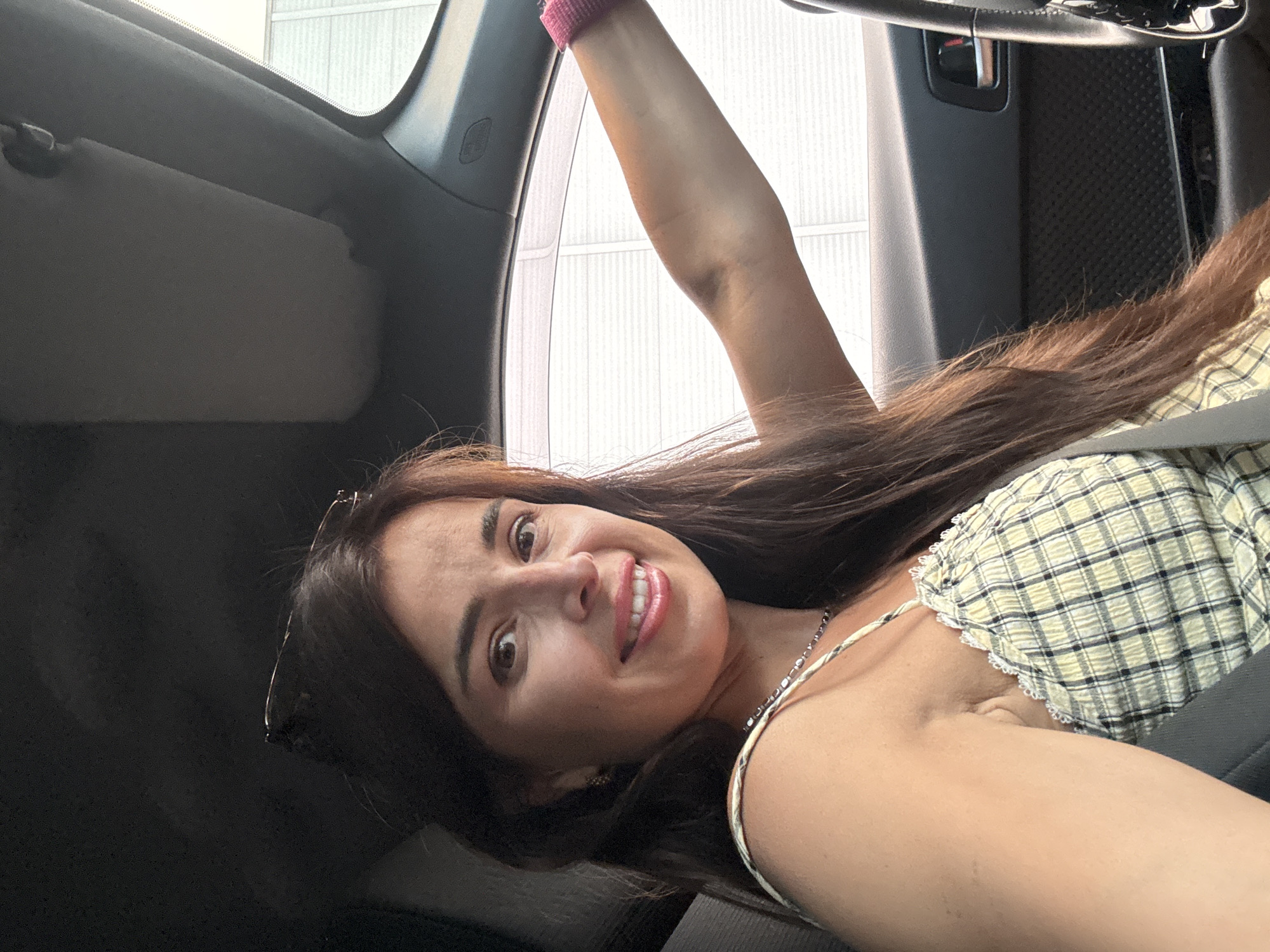 A woman is in the driver's seat of a car. She has long, dark brown hair and is wearing a yellow plaid top with white trim. She is smiling and looking at the camera. Her left arm is resting on the steering wheel, and she is wearing a pink bracelet. The car's interior is visible, including the dashboard and a sun visor. The background outside the car appears to be a building with horizontal white siding.