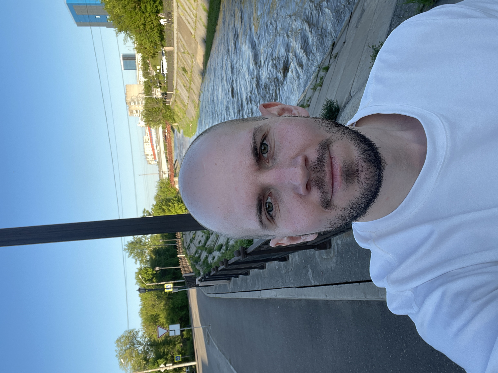 The image is a selfie of a light-skinned man with a shaved head and a dark beard, wearing a white hooded sweatshirt. He is standing near a river in Krasnoyarsk, Russia, as evidenced by the presence of the Yenisey River embankment with its characteristic reinforced concrete channel and architectural elements. In the background, there is urban development, including buildings of various heights and design. In the sky, there are visible electric wires. On the side of the photo, there's an adjacent road with street signs and a pedestrian crossing. The photo appears to be taken during the daytime under clear conditions.