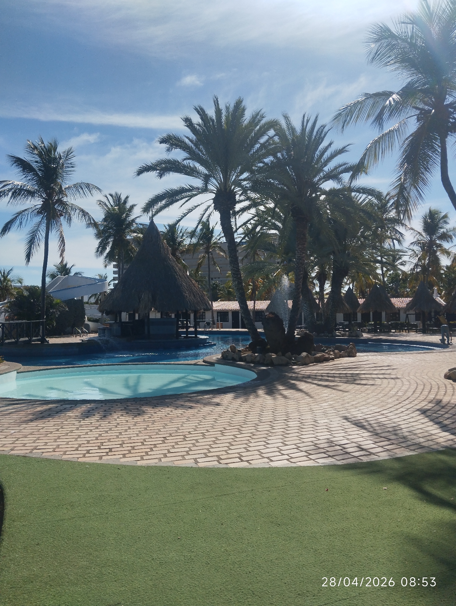 This sunlit image depicts a serene tropical resort pool area in the early morning. The scene features multiple tall palm trees with green fronds, casting long, sharp shadows across the ground, indicating bright sunshine. The sky is a clear blue with some faint, wispy white clouds.
In the foreground, there's a small, circular swimming pool with light blue water. This pool is connected to or adjacent to a larger, irregularly shaped main pool. A notable feature within the larger pool is a swim-up bar sheltered by a large, conical thatched-roof hut, with water flowing around it. Other smaller thatched-roof huts, possibly cabanas or shaded seating areas, are visible further back around the pool deck. A decorative rock formation with a water fountain adds to the aesthetic appeal of the main pool.
The pools are surrounded by a patterned paved surface, which transitions into an area of artificial green grass in the lower part of the frame. In the background, behind the dense palm foliage, resort buildings or other structures can be seen.
No people are visible in the image, contributing to a peaceful and inviting atmosphere that suggests a tranquil start to the day. The clear skies and bright sunlight indicate good weather. A timestamp "28/04/2026 08:53" is visible in the bottom right corner, confirming the morning time. The overall impression is that of a warm, relaxing vacation destination.