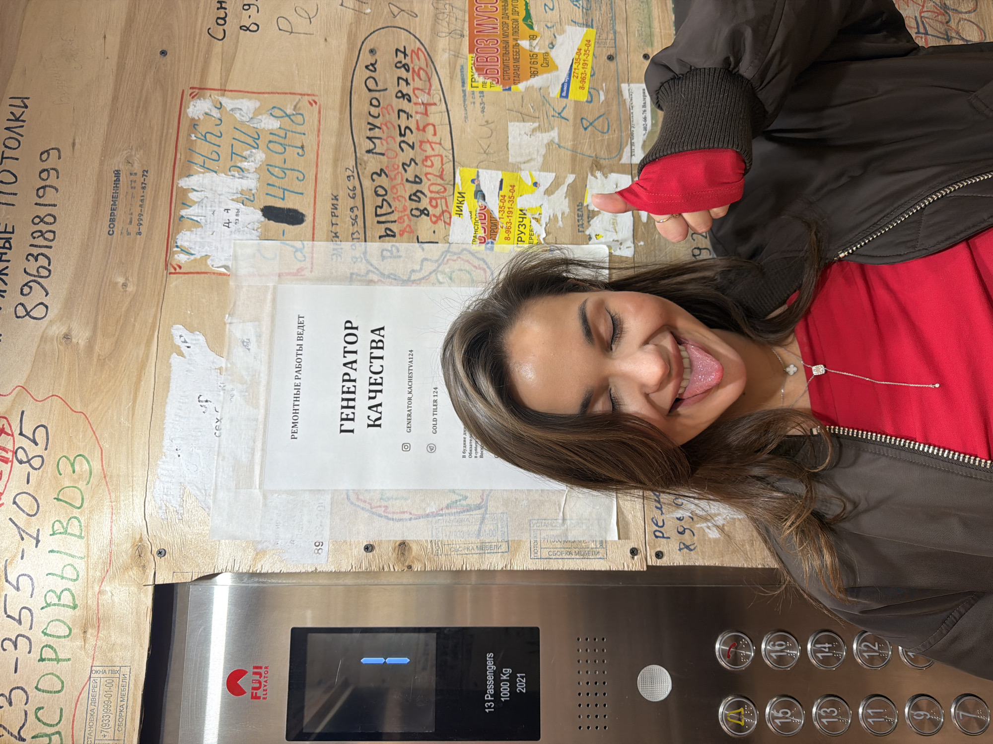 The image shows a young woman in an elevator, giving a thumbs up, with her tongue sticking out. She wears a red shirt and a black jacket, and a silver necklace. The elevator appears to be of the brand "FUJI Elevator" and manufactured in 2021. It can carry 13 passengers with a maximum weight of 1000 kg. The interior of the elevator is covered in various advertisements and phone numbers, typical for the city of Krasnoyarsk, Russia. These advertisements appear to be related to services such as junk removal, furniture assembly, and ceiling installation. Some numbers include 89631881999, 23-355-10-85, and 8-963-257-8787.