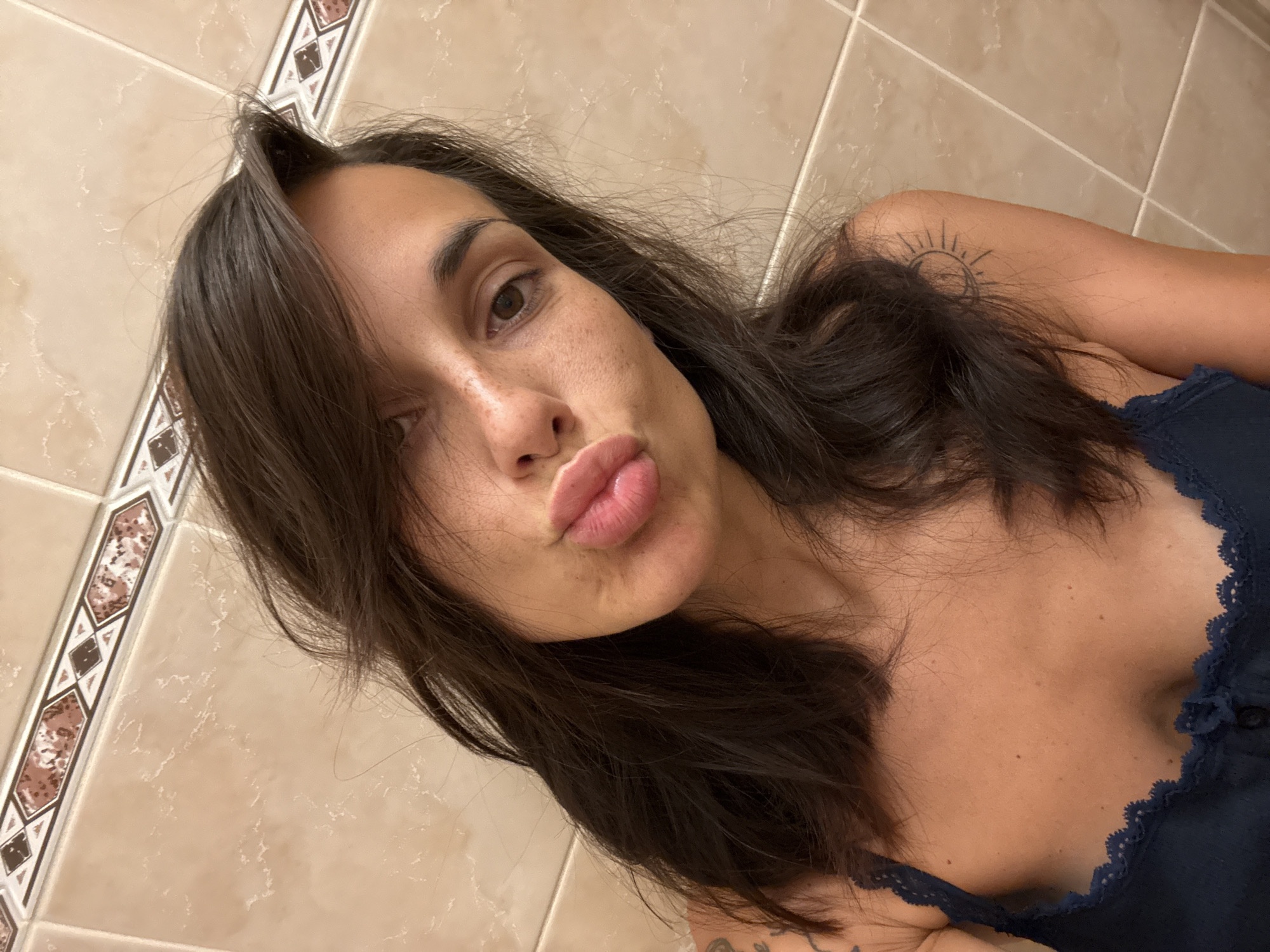A woman is taking a selfie in a bathroom with tiled walls. She has dark hair and is making a kissing face. She is wearing a dark blue camisole. There are tattoos on her shoulder and arm.