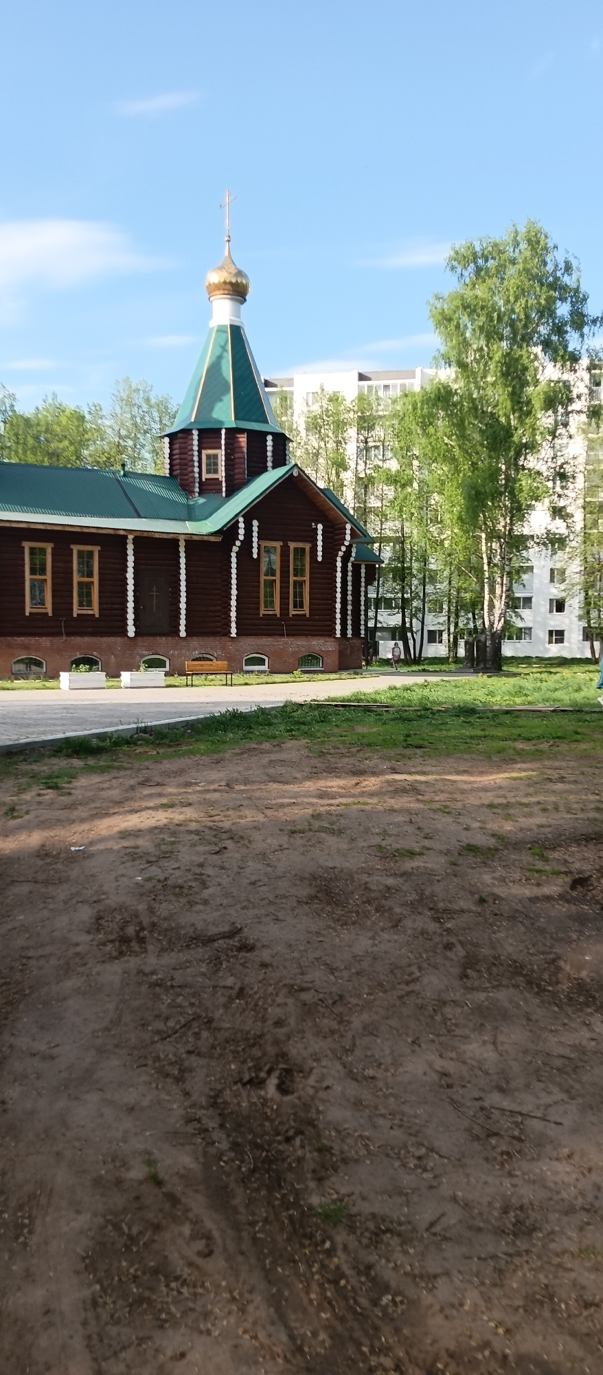 The image displays a wooden Orthodox church situated in what appears to be a park-like setting, with modern urban buildings in the background. The church has a dark brown, log-cabin-style exterior with decorative white vertical accents and light-framed rectangular windows. Its roof is green, culminating in a steeple topped with a golden onion dome and an Orthodox cross. The foundation of the church appears to be made of brick.
In the immediate foreground, a large area of bare, light brown earth is visible, with sparse patches of grass. A paved path or sidewalk runs in front of the church, where a wooden bench and two white planters are placed. Beyond the church, several tall, leafy green trees, likely birches, stand, partially obscuring a multi-story white apartment building. A single person is faintly discernible in the mid-ground near the apartment building, on the right side of the image.
The weather is bright and clear, with a blue sky and a few faint clouds, suggesting it is daytime, possibly morning or late afternoon given the soft lighting. No specific activity or event is taking place, and no discernible text is visible in the image. The scene offers a peaceful contrast between traditional religious architecture and modern residential structures.