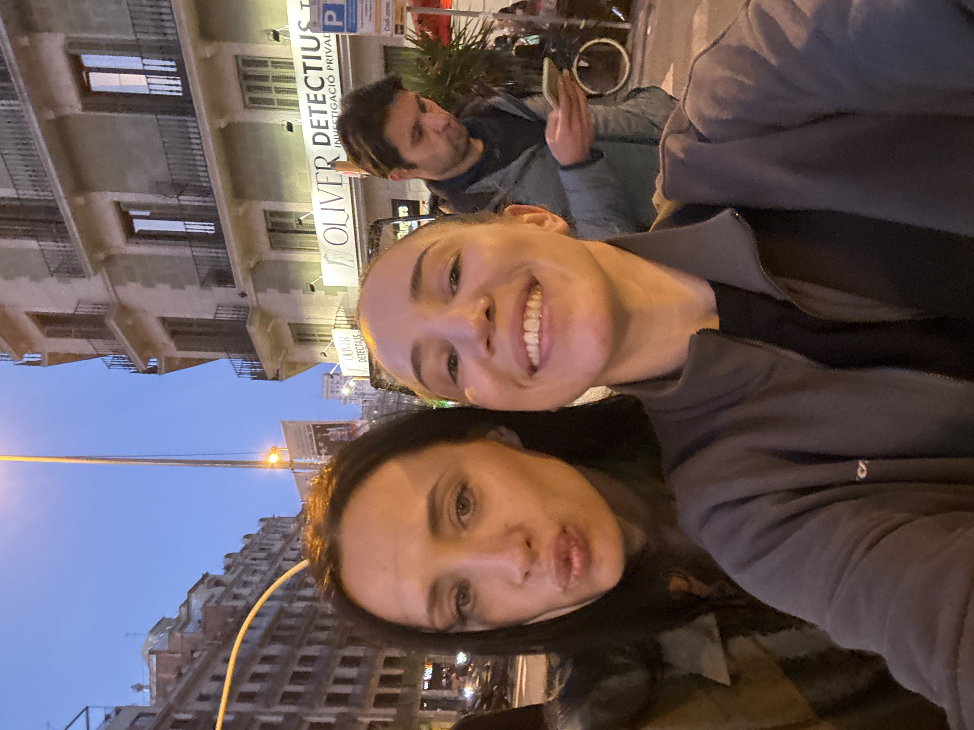 The image shows two women in a selfie. One woman is smiling, and the other is pouting. In the background, a man is looking at his phone. A sign reads "OLIVER DETECTIUS INVESTIGACIO PRIVADA". The building facade appears to be a typical older style in Barcelona, Spain.