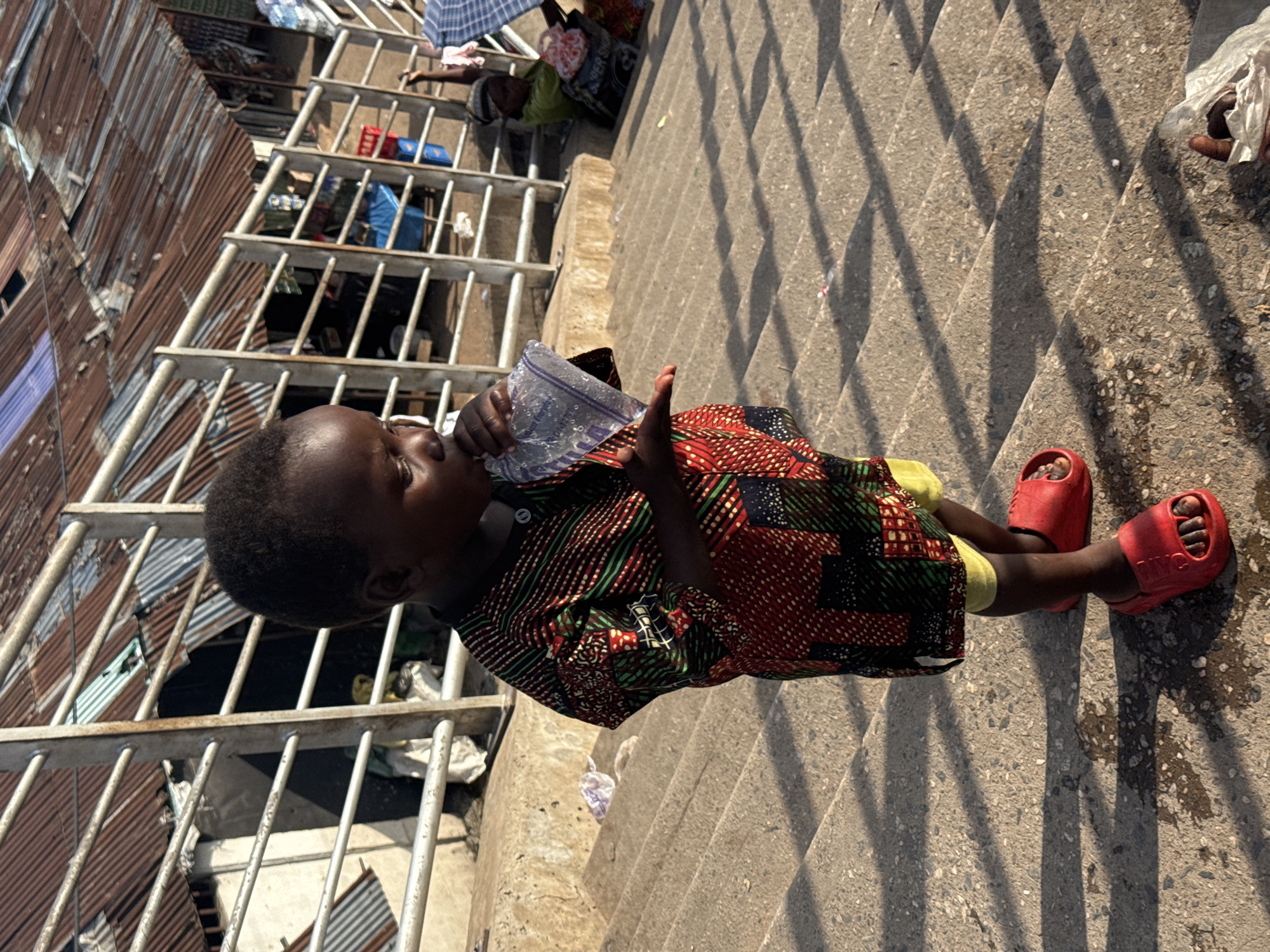 The image shows a young child sitting on concrete stairs, holding a plastic bag with water. The child is wearing a colorful patterned shirt and yellow shorts, with red sandals on their feet. Behind the child, there are corrugated metal structures and what appear to be makeshift shelters with metal railings. In the background, other people can be seen, some sitting or lying down, and a blue umbrella is also visible. The scene is lit by sunlight, casting distinct shadows from the railings onto the stairs, suggesting it is daytime. The setting appears to be a densely populated, possibly informal settlement. The child's expression is neutral as they drink from the bag. There is no visible text in the image.