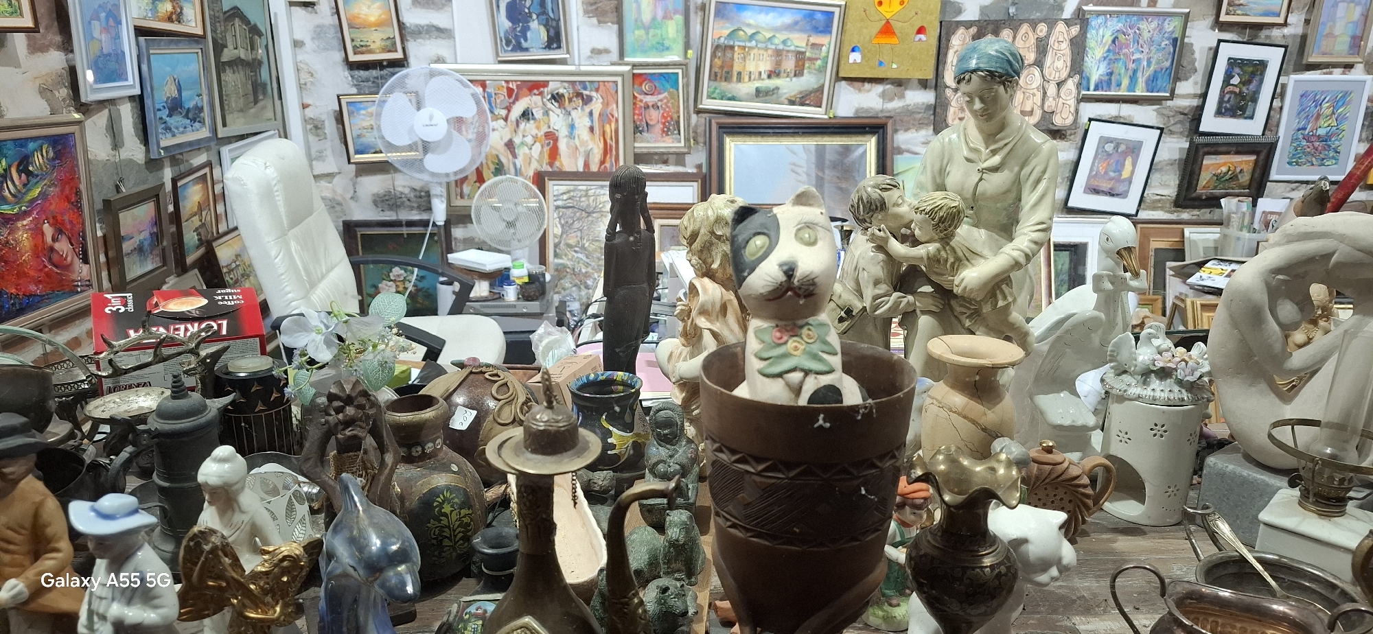 The image depicts an indoor market stall in Yambol, Bulgaria, filled with an eclectic assortment of decorative items. The stall is densely packed with various objects, primarily figurines and art pieces, arranged on shelves and tables. In the foreground, a ceramic cat figurine sits in a dark, textured vase. Behind it, a large cream-colored sculpture of a woman cradling two children is prominently displayed.
The walls are adorned with numerous framed paintings and artworks, showcasing diverse styles and subjects, including landscapes, abstract pieces, and figurative compositions. Several electric fans are visible, suggesting a warm environment. Other items scattered throughout the stall include ceramic vases, metal decorative pieces, small statues of animals and human figures, and various decorative trinkets. The overall impression is one of a cluttered but visually interesting space, likely a shop specializing in antiques or handcrafted decorative items. The presence of a mobile phone watermark in the lower left corner indicates the image was captured by a modern device.