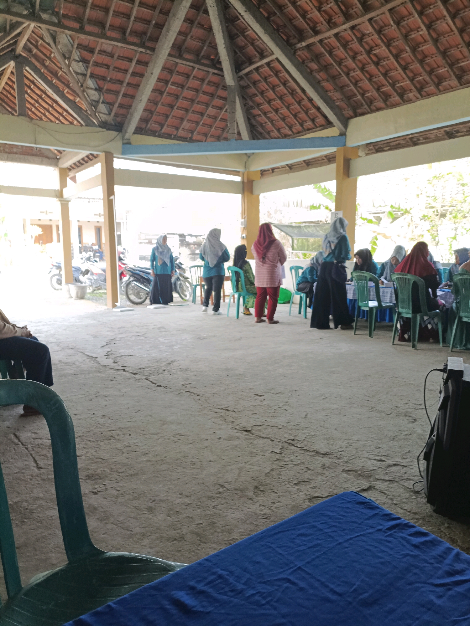 The image depicts an outdoor gathering or event under a covered structure with a tiled roof supported by wooden beams and pillars. The floor is concrete, showing some cracks and wear. In the background, there are buildings and vegetation. Several people are present, mostly women wearing hijabs and modest clothing. Some are standing, while others are seated at tables. There are several motorcycles parked in the background. The scene appears to be in an open-air area, possibly a community space or market in Rancang Kencono, Indonesia. The lighting suggests it is daytime, with sunlight filtering through the open sides of the structure. A green plastic chair is visible in the foreground, and a blue cloth covers a surface in the lower right quadrant of the image. A black speaker is also visible on the right side.