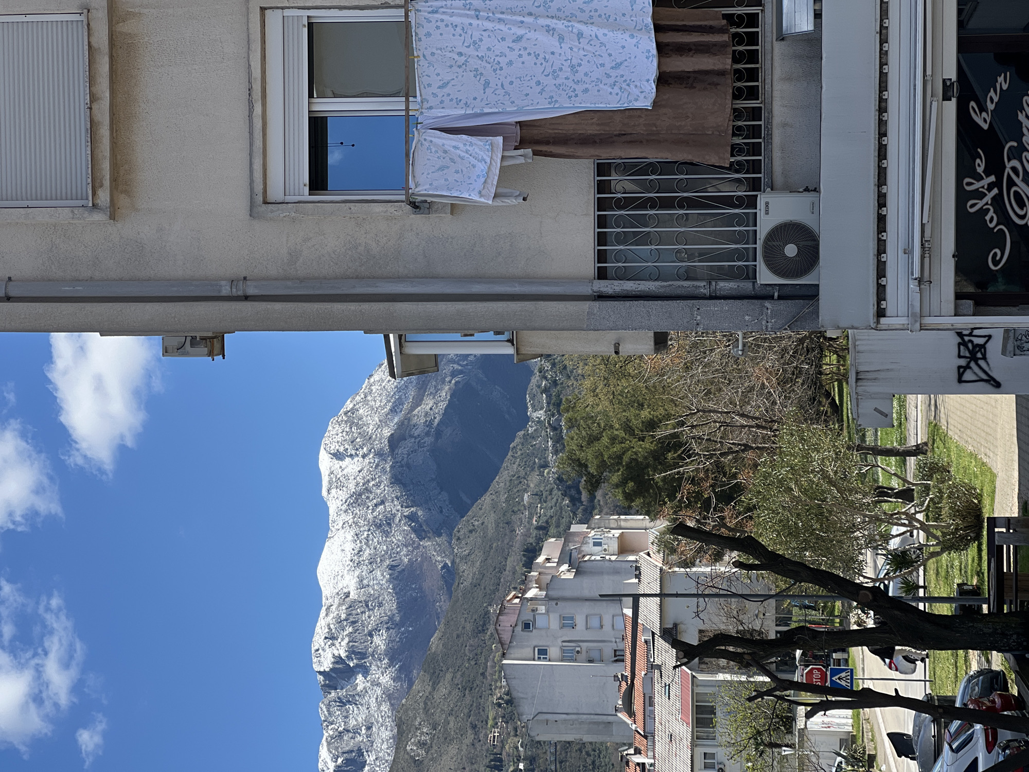 The image depicts a street scene in Bar, Montenegro, with a view of a snow-capped mountain in the background. In the foreground, there are buildings with clothes hanging out to dry from windows. A "Cafe Bar" sign is visible on one building. The street is lined with trees and parked cars. There is a traffic sign visible in the lower right corner of the image. The sky is clear and blue with some clouds.