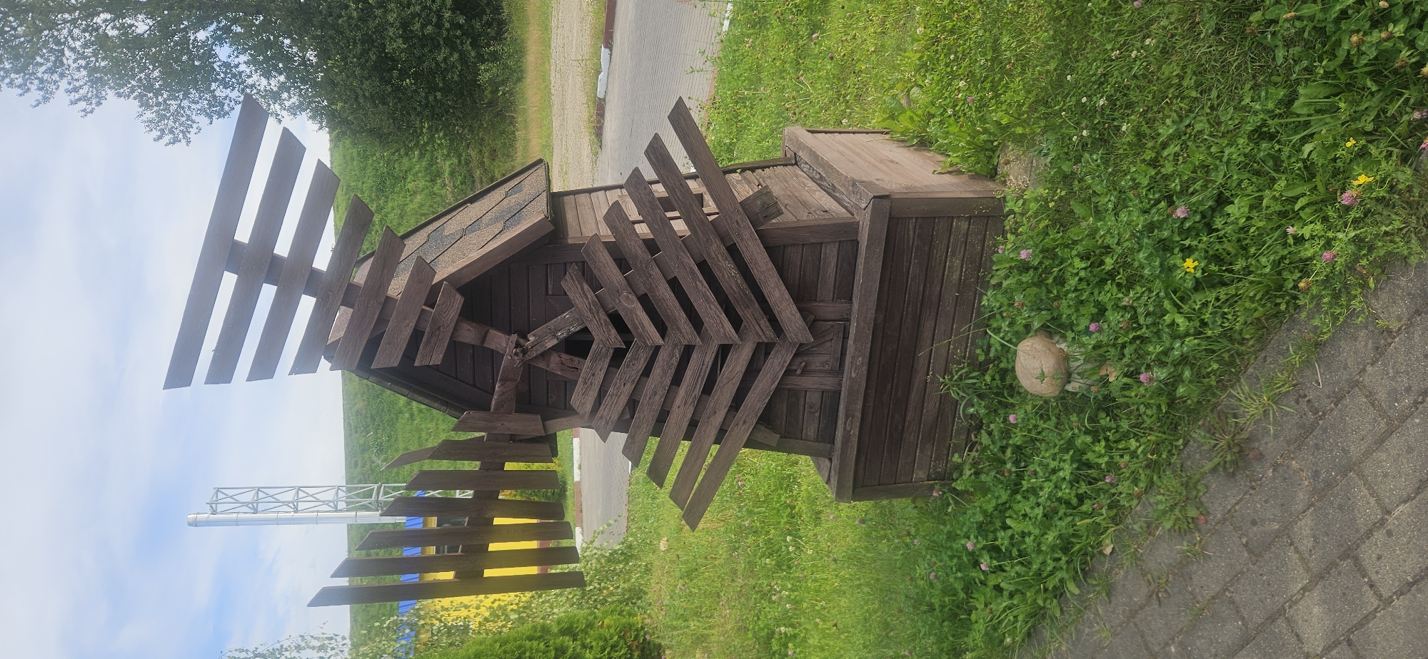 In Vitsebsk, Belarus, a small, rustic wooden windmill sculpture stands amidst lush green grass and clover. The windmill, with its weathered brown planks, features a distinctly abstract design, especially in its fan-like blades which are angled haphazardly. A dark, shingled roof tops the structure. At the base of the windmill, a small, mushroom-shaped garden ornament is partially visible amongst the foliage. To the left, a portion of a paved walkway is visible, made of grey rectangular bricks. Beyond the windmill, a tall, silver metallic structure, possibly a chimney or industrial vent, rises against a pale blue sky with scattered clouds. A yellow and blue fence stands behind the windmill and to the left, partially obscured by green bushes and trees. The environment appears to be an outdoor public space, possibly a park or recreational area, under daylight conditions.