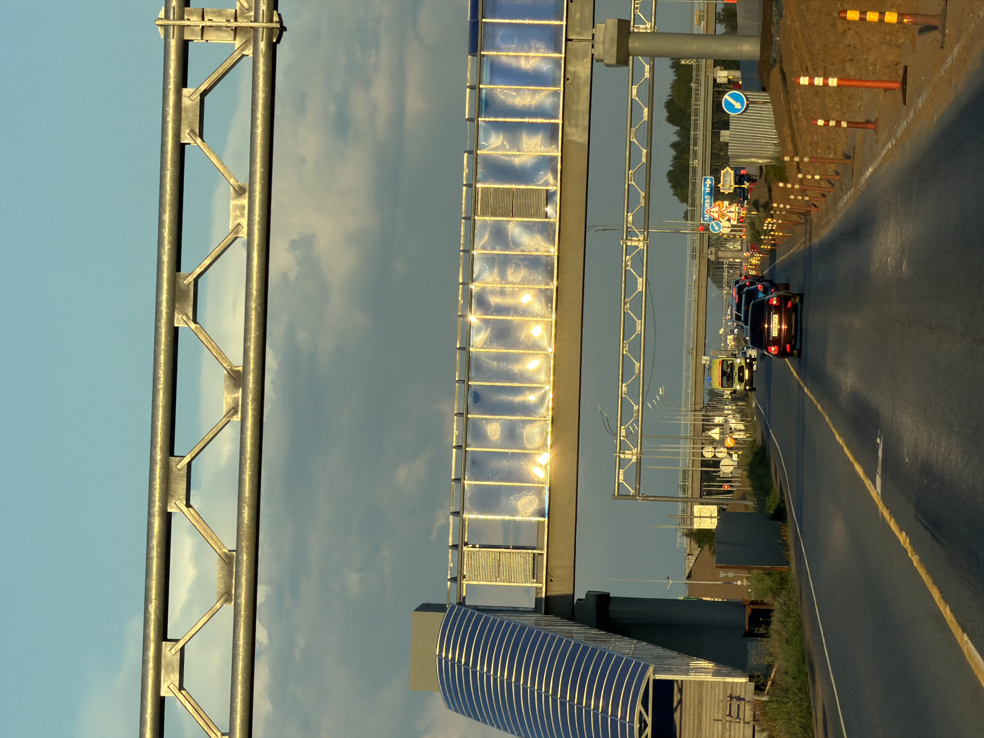 The image depicts a highway scene in Derzhavino, Russia, during the late afternoon or early evening, indicated by the warm, angled sunlight. A multi-lane paved road dominates the foreground, with a yellow line marking the center. Several vehicles are driving on the road, including a dark-colored sedan in the immediate foreground and a yellow ambulance or emergency vehicle further ahead, signaling a potential departure from the scene.
Overhead structures are prominent. A large, metallic truss system spans the width of the image at the top, with a suspended pedestrian walkway or overpass running parallel to the road below it. This walkway features a translucent, rippled blue and reflective covering, with internal lights creating bright reflections. To the left of the walkway, a ramp-like structure with a similar blue, corrugated covering leads up to it.
The roadside is lined with orange traffic cones and posts, some with reflective white and orange stripes, suggesting ongoing roadwork or traffic management. Beyond the road, there are various traffic signs, some directing to "ДомОВО 16к" (Domovo 16k), indicating a destination. A glimpse of trees and a distant horizon suggests the road is on the outskirts of the urban area. The sky is a clear blue with scattered, wispy clouds, catching the golden light of the setting sun. The overall environment appears to be a functional infrastructure area, likely part of a highway or major arterial route within the city.