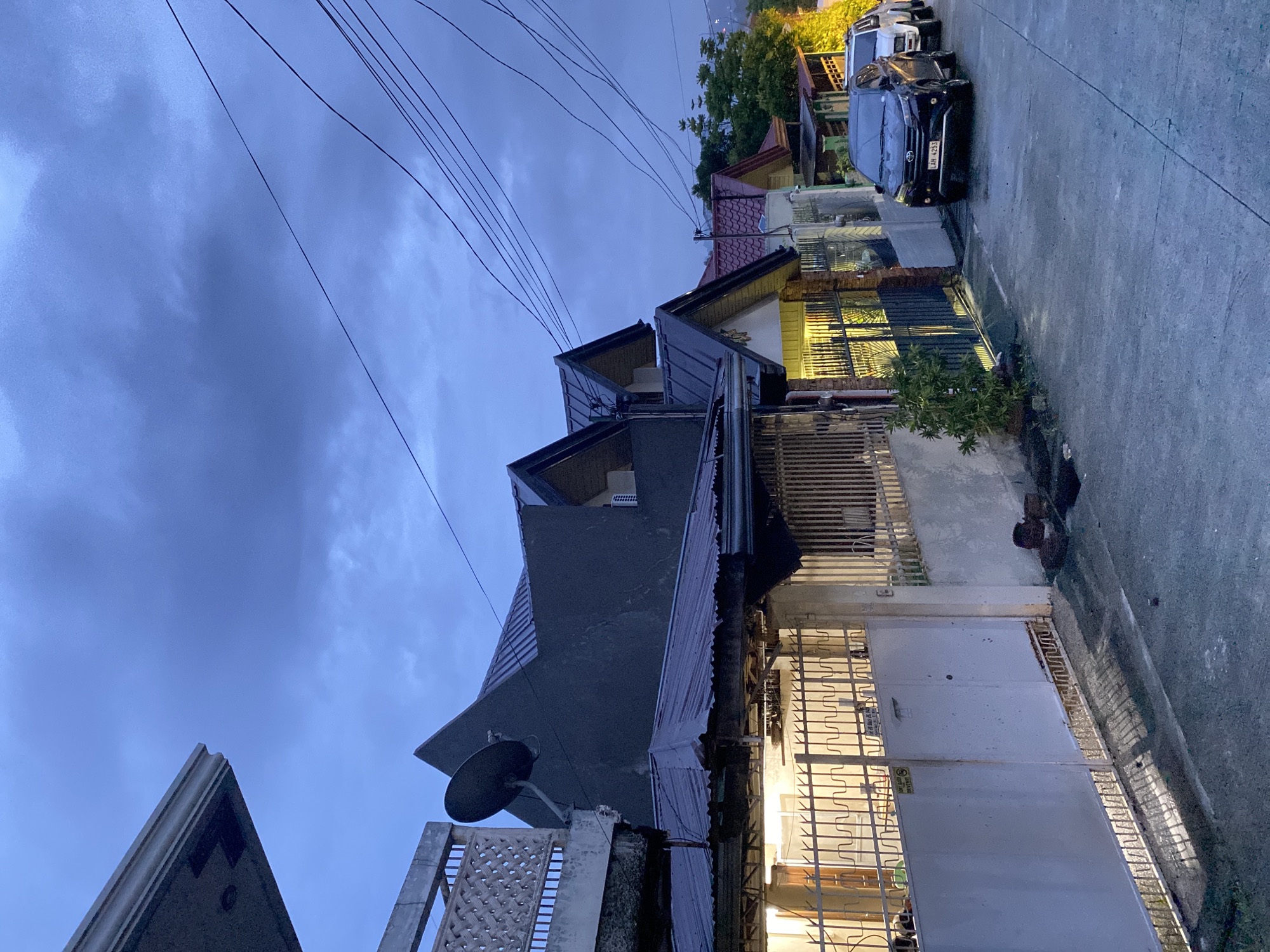 This is an outdoor, eye-level shot of a street scene in Davao City, Philippines, taken at dusk. The sky is a deep, cloudy blue, with several black electrical wires crisscrossing the upper portion of the frame. In the foreground, a gray residential building with a dark roof and angled windows dominates the left side of the image. To the right of this building, a smaller house with a red-tiled roof and yellow siding is visible, with interior lights glowing warmly from behind barred windows and a metal gate. Beyond this, a street runs along the right side of the image, bordered by more houses and greenery. Two cars are parked on the street: a black sedan in the middle ground, and a white vehicle further down the street. The overall mood is calm and quiet, with the transition from day to night evident in the lighting and sky. A white metal gate with a decorative pattern is closed across the entrance to one of the properties. Text is visible on a sign attached to the white gate, reading "DO NOT LEAVE ANYTHING OUTSIDE."