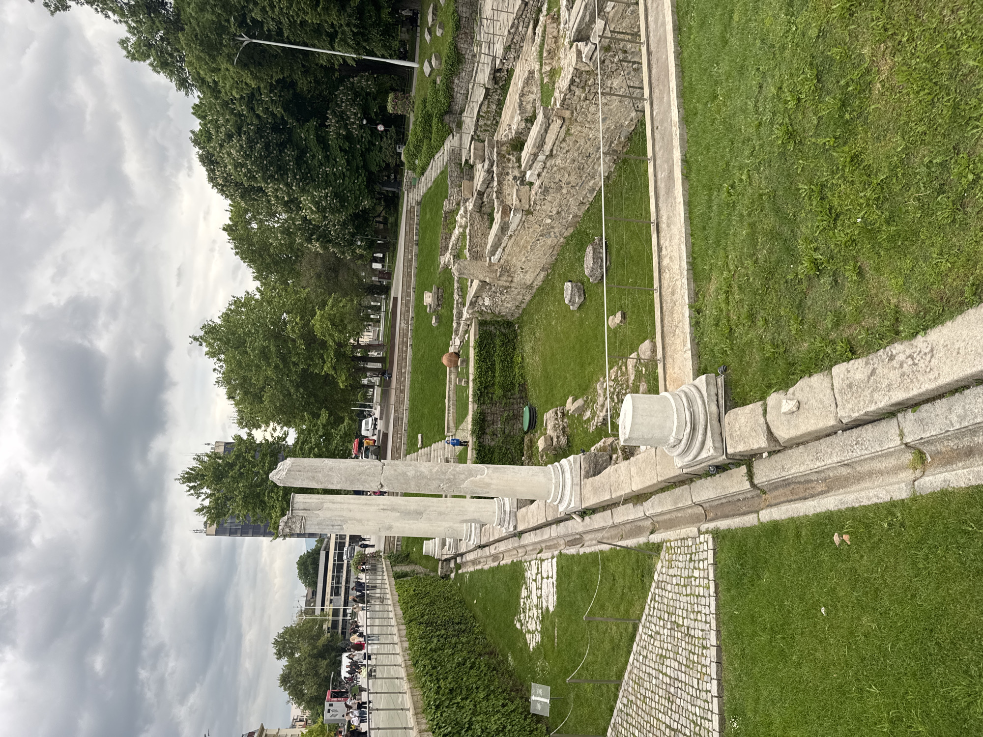 The image shows the Ancient Stadium of Philippopolis in Plovdiv, Bulgaria. The scene is captured from an elevated position, overlooking the remnants of the stadium. Several stone pillars are visible in the foreground, leading into the excavated remains of the stadium. Grass surrounds the ruins, which are enclosed by a low fence. Trees are in the background, partially obscuring buildings in the distance. People are present along the street, suggesting a bustling city environment. The sky is overcast with clouds.