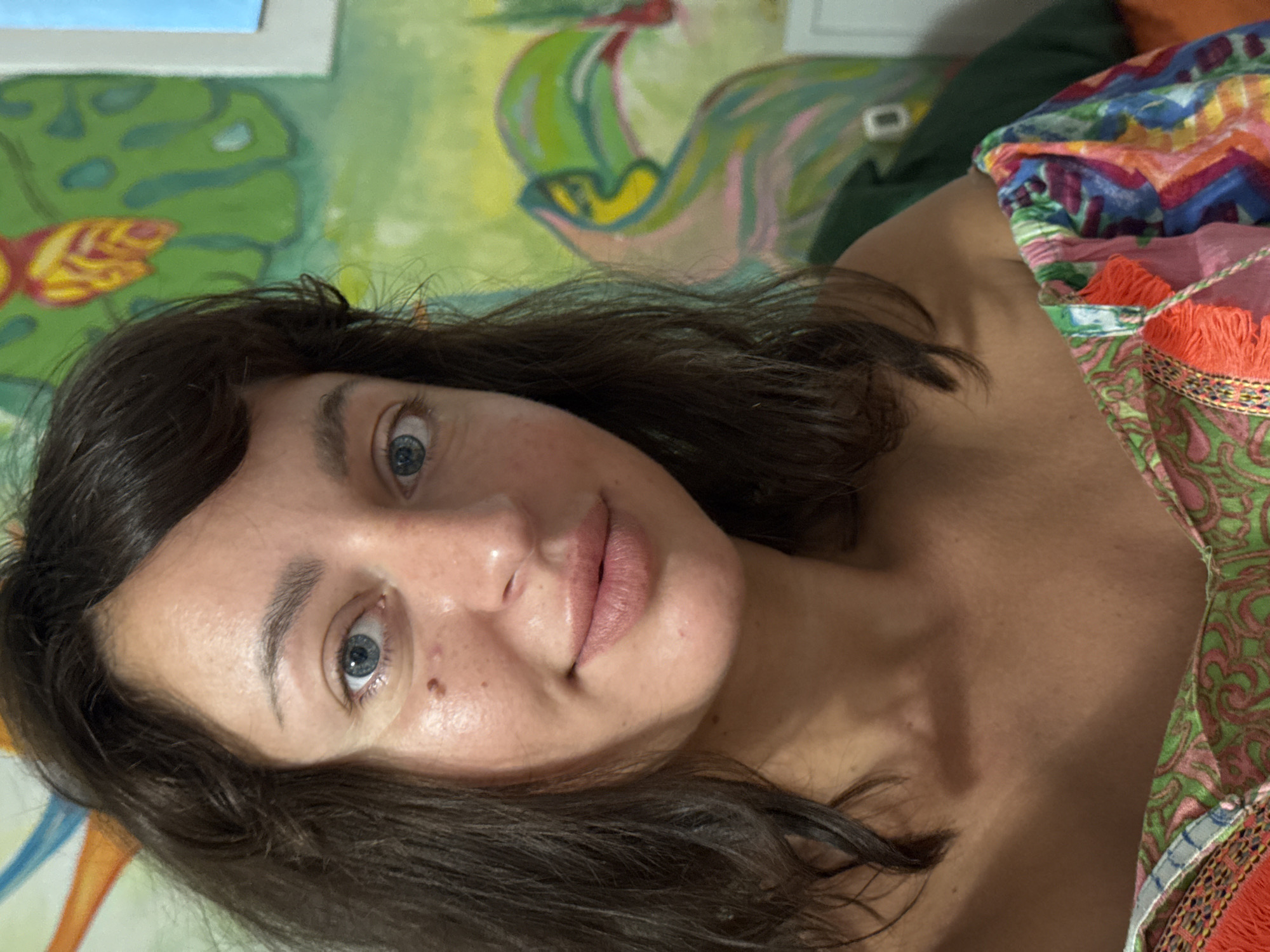 This is a close-up, low-angle selfie of a woman with dark brown hair. She is wearing a colorful, patterned garment with orange fringe. The background appears to be a wall with a colorful mural featuring green and yellow organic shapes and a blue element at the top left. The woman's eyes are open and looking towards the camera. Her lips are slightly parted, and she has fair skin with some freckles and a mole on her cheek. The lighting suggests it could be daytime, though no specific time or weather conditions are evident. The image was taken in San Benedetto del Tronto, Italy. No visible text is present in the image.